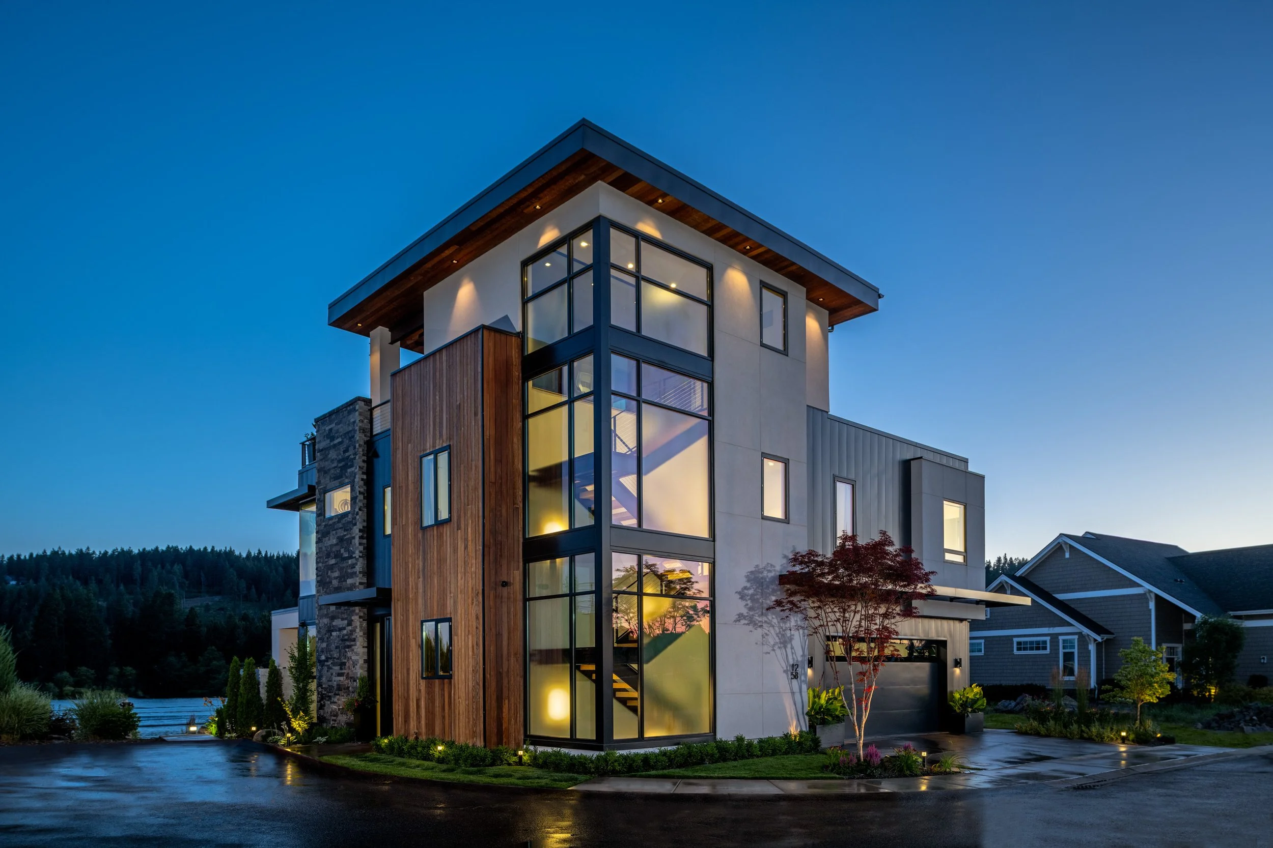 Modern multi-story house with large glass windows, wooden and stone exterior, surrounded by landscaping at dusk.