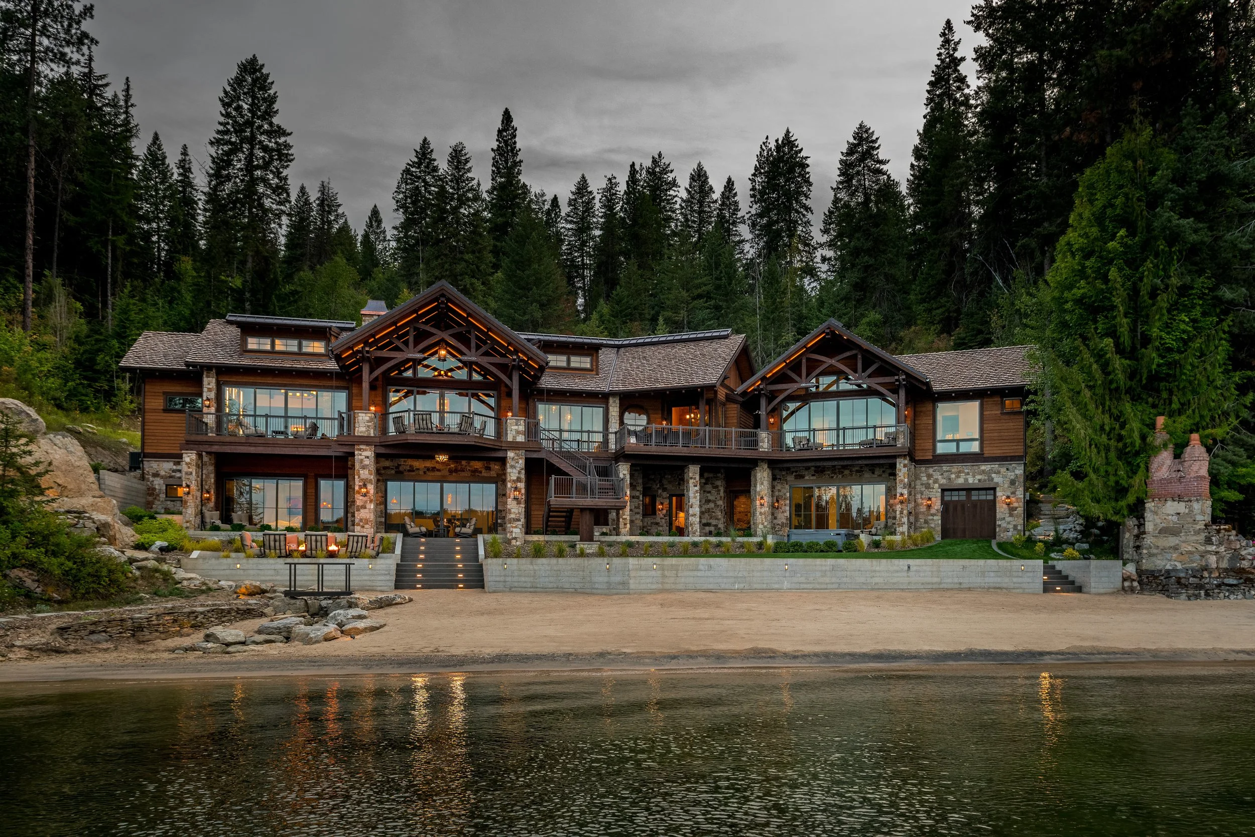 Luxury house with stone and wood exterior, large windows, two upper balconies, located on a beach with trees and mountains in the background, reflecting in the water.