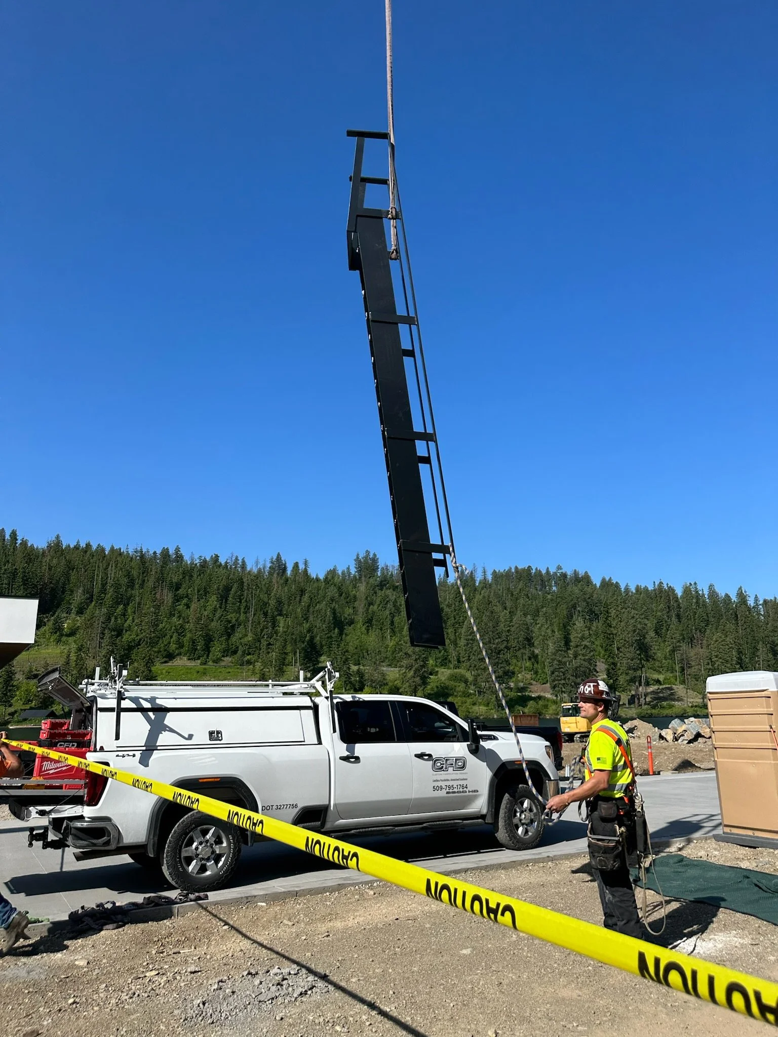Construction worker standing near a white truck with a large black sign or panel hanging above, secured by ropes. The area is cordoned off with yellow caution tape, and there are construction materials and equipment nearby. The background features a forested hillside under a clear blue sky.