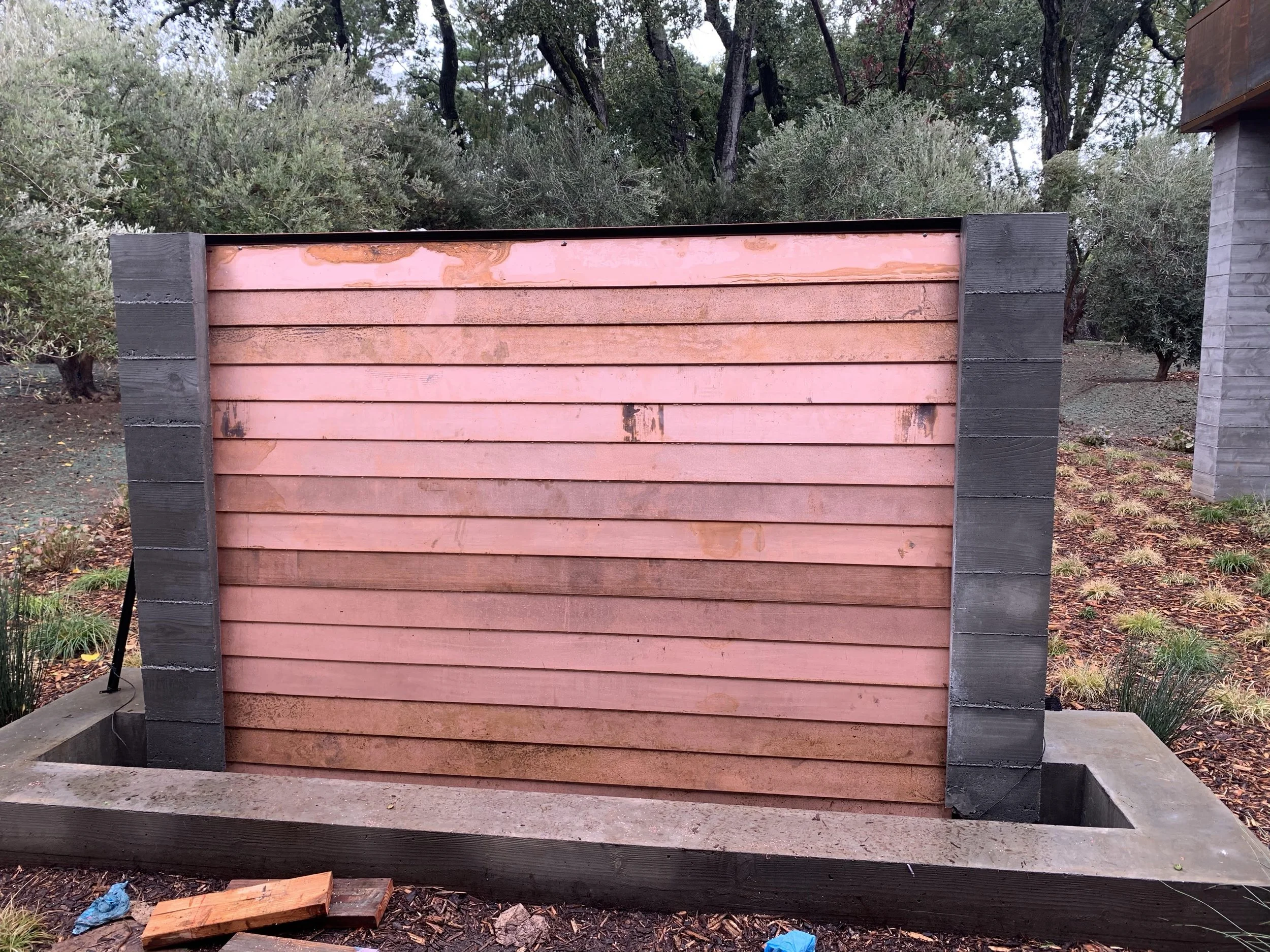 A half-finished outdoor water feature with wooden slats and a concrete base, surrounded by trees and plants.