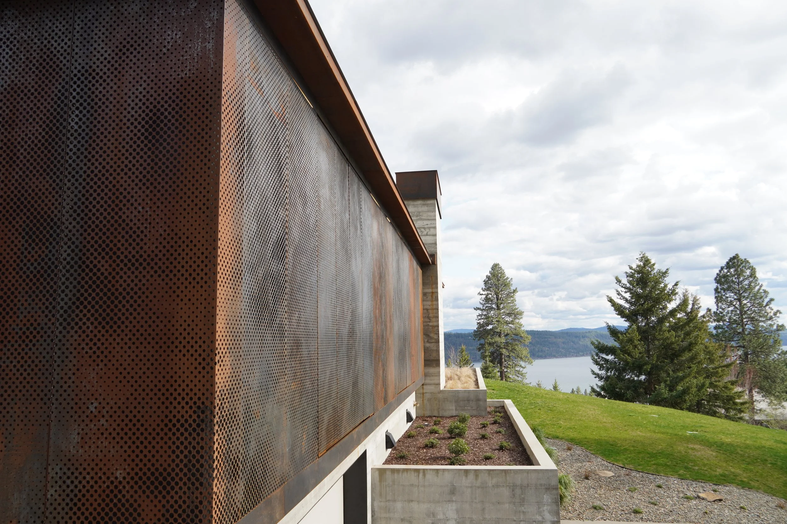 Side view of a modern building with perforated metal panels, overlooking trees, a body of water, and mountains under a cloudy sky.