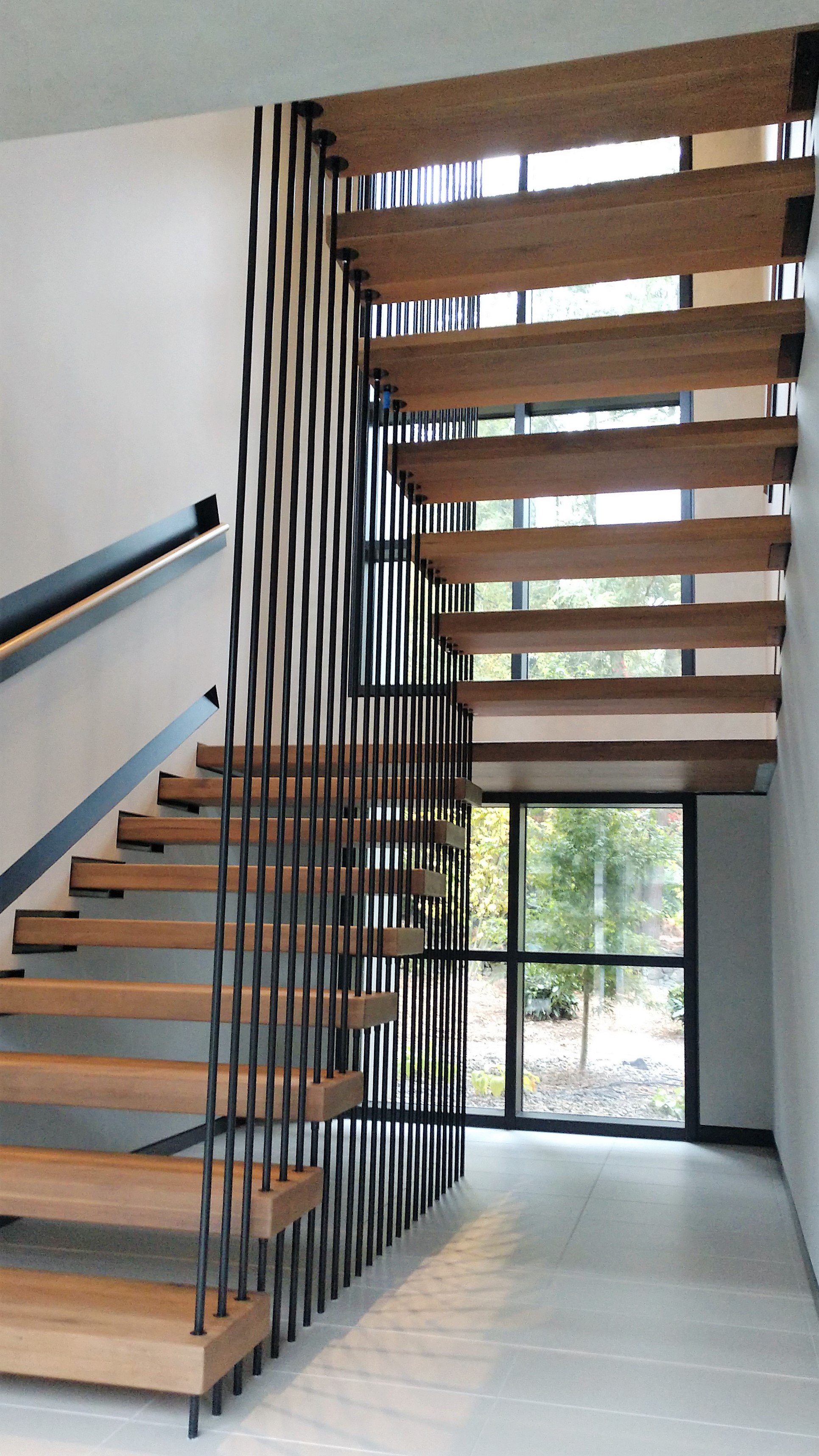 Modern interior staircase with wooden steps and black vertical railing, large windows in the background with trees outside.