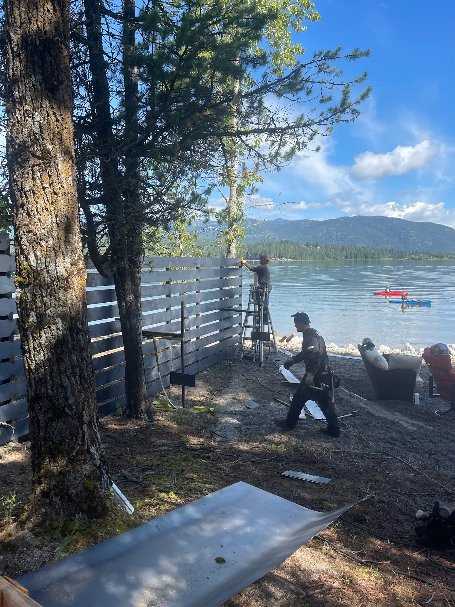 Two workers installing a wooden fence along the lakeshore, with trees and mountains in the background, and kayaks floating on the water.