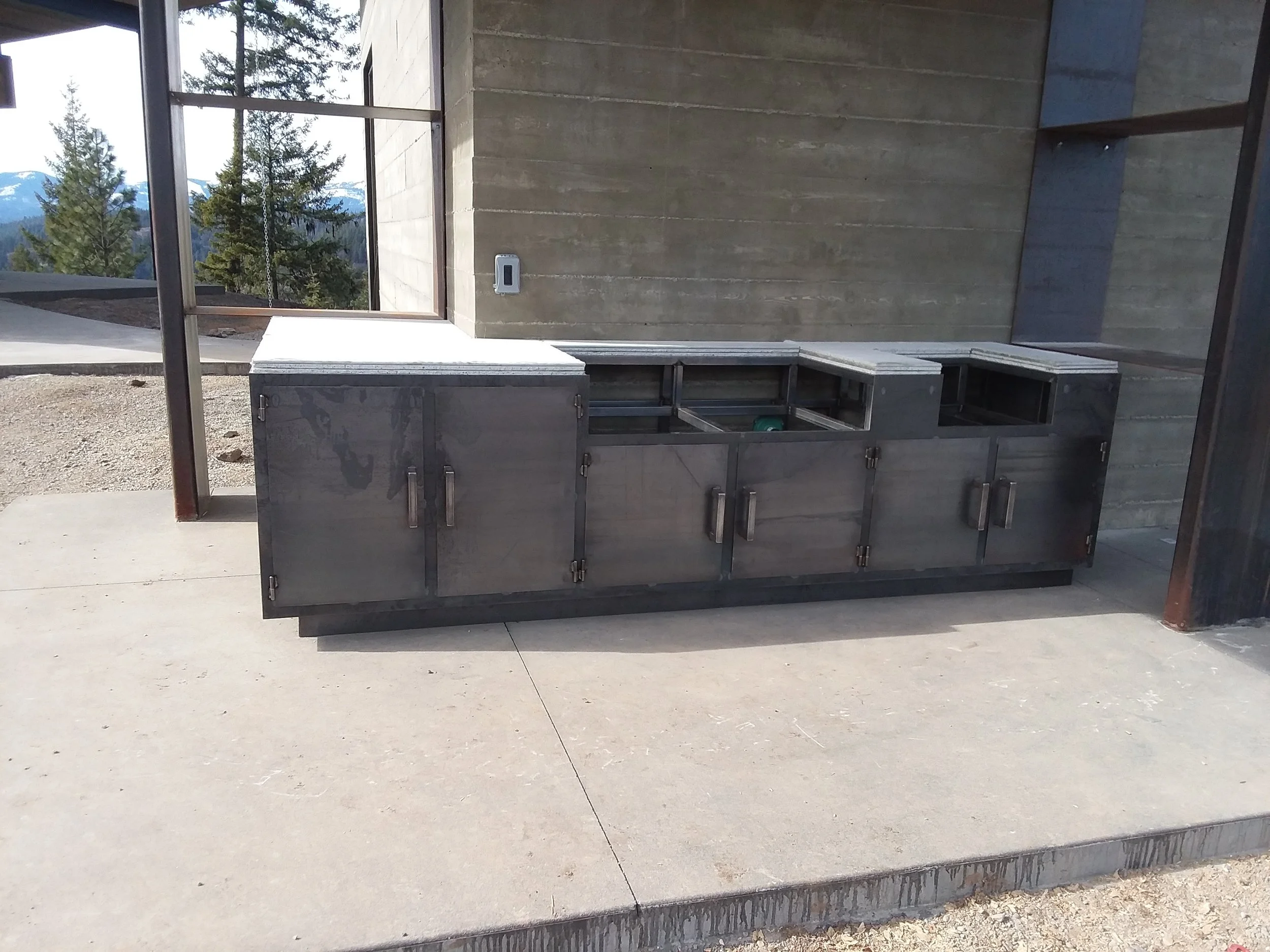 Empty outdoor kitchen cabinet with stainless steel doors on concrete patio, against a modern building with large windows and a mountain landscape in the background.
