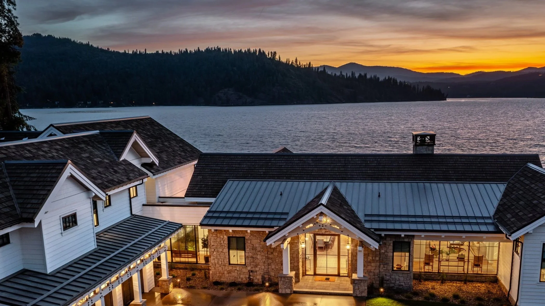 A lakeside house at sunset with a view of the water and distant hills, featuring a modern design with large windows and a stone and white exterior.