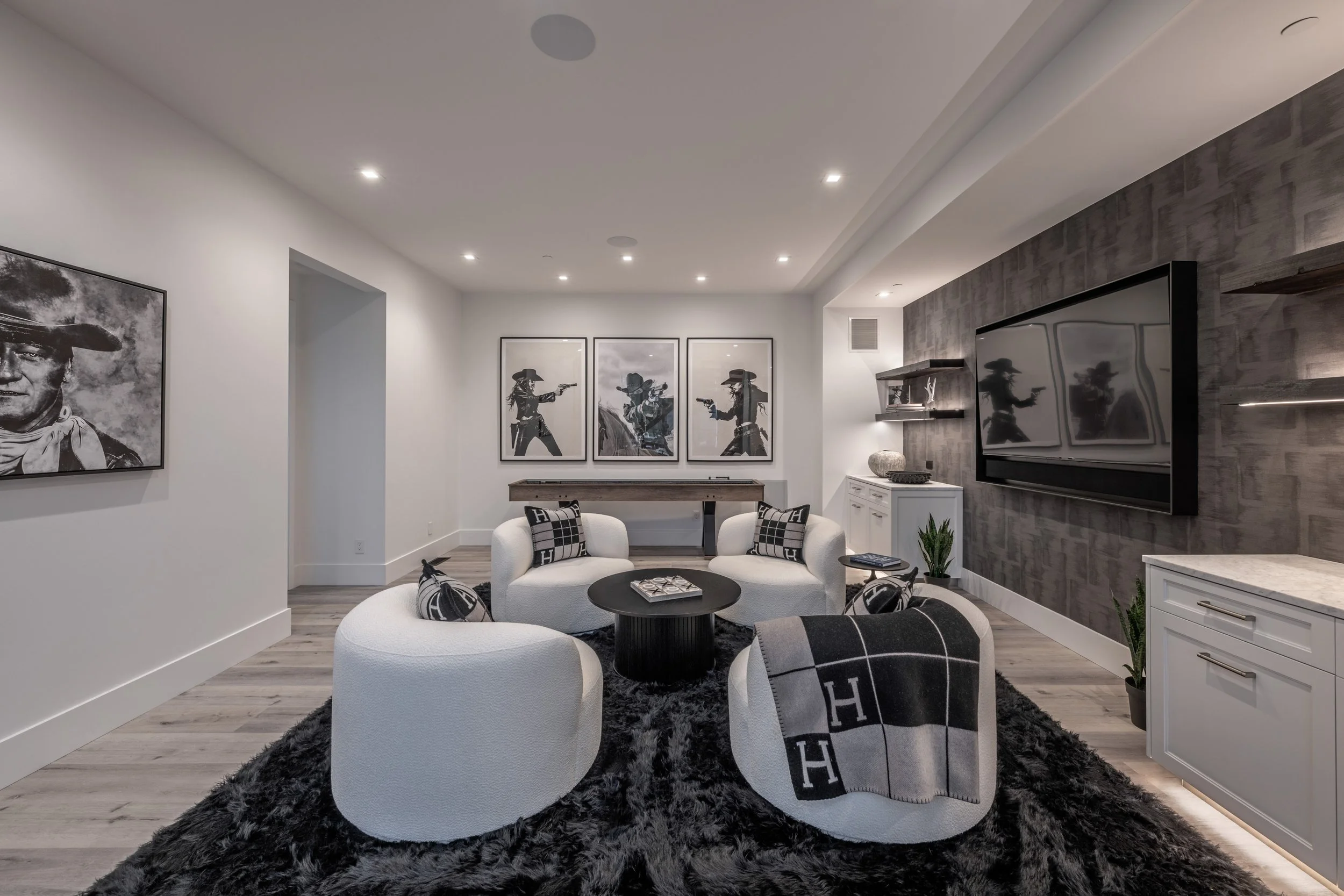 Modern living room with white curved sofas, black circular coffee table, black and white pillows, gray plush rug, wall-mounted TV, black wood paneled accent wall, art photos of cowboys, white cabinets with marble countertops, potted plants, and decor