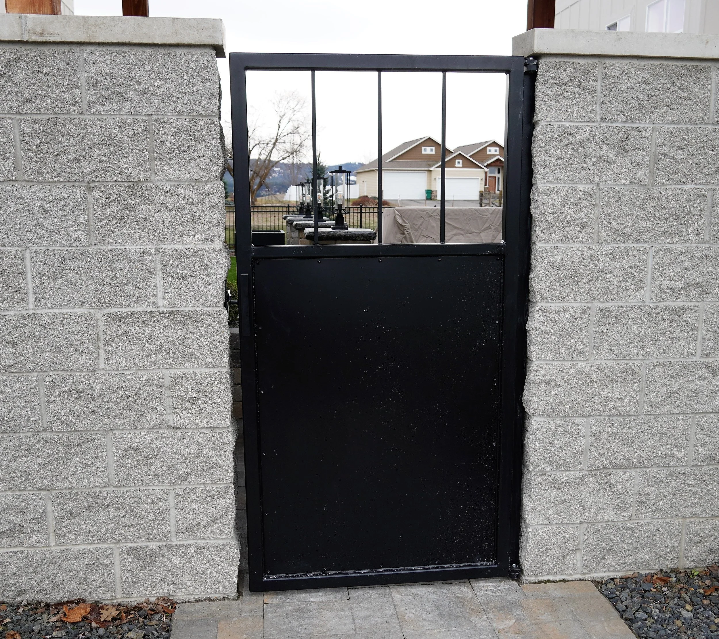A black metal gate with barred top panel, set between two concrete block walls, with a residential neighborhood in the background.