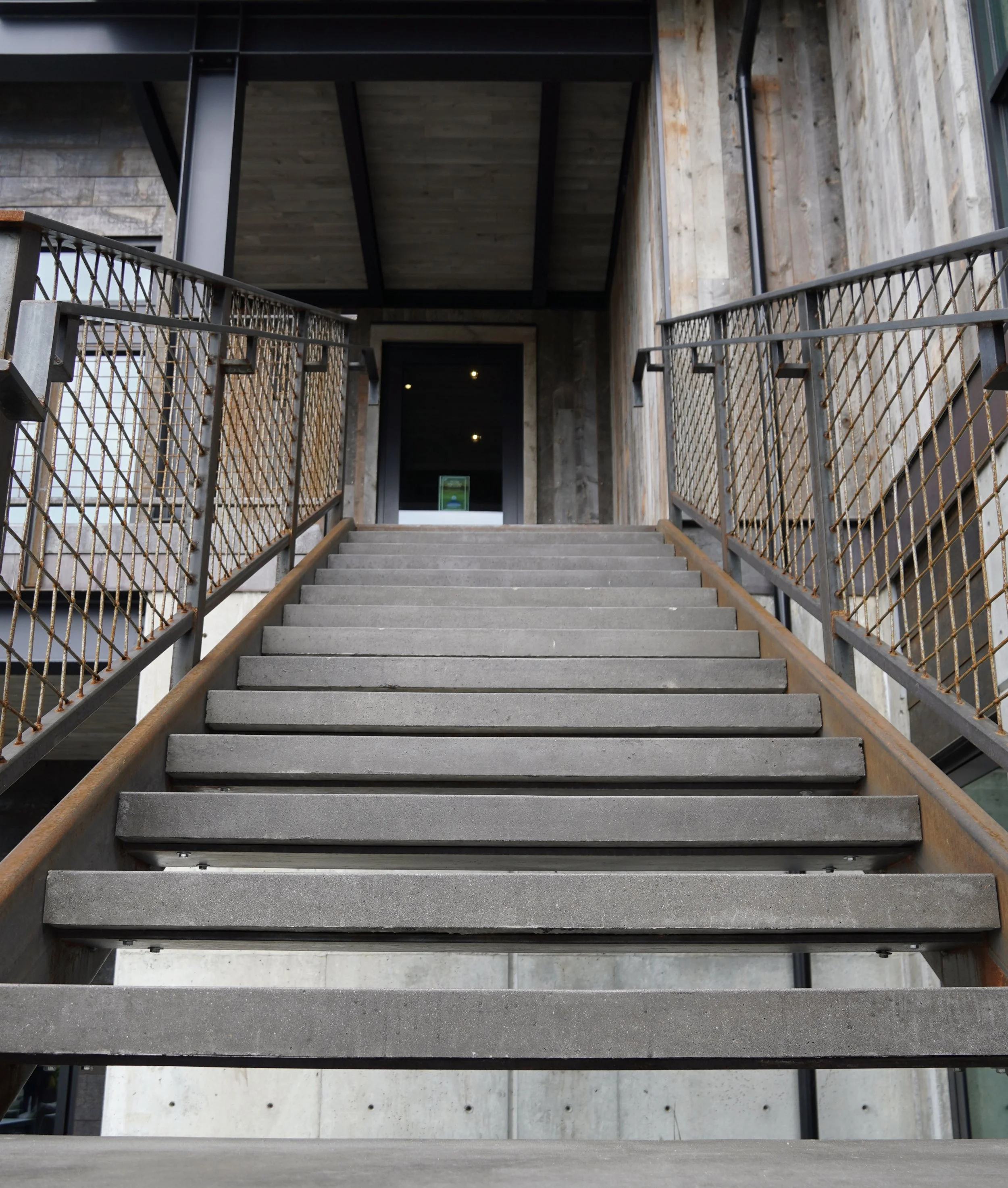 Concrete staircase with metal railings leading up to a door in a modern building with wooden and concrete exterior.