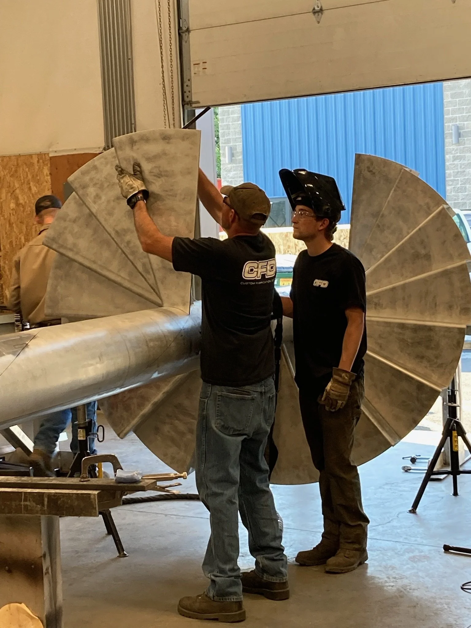 Two workers in black shirts with 'CFD' logo assembling a large, metallic turbine blade inside a workshop.