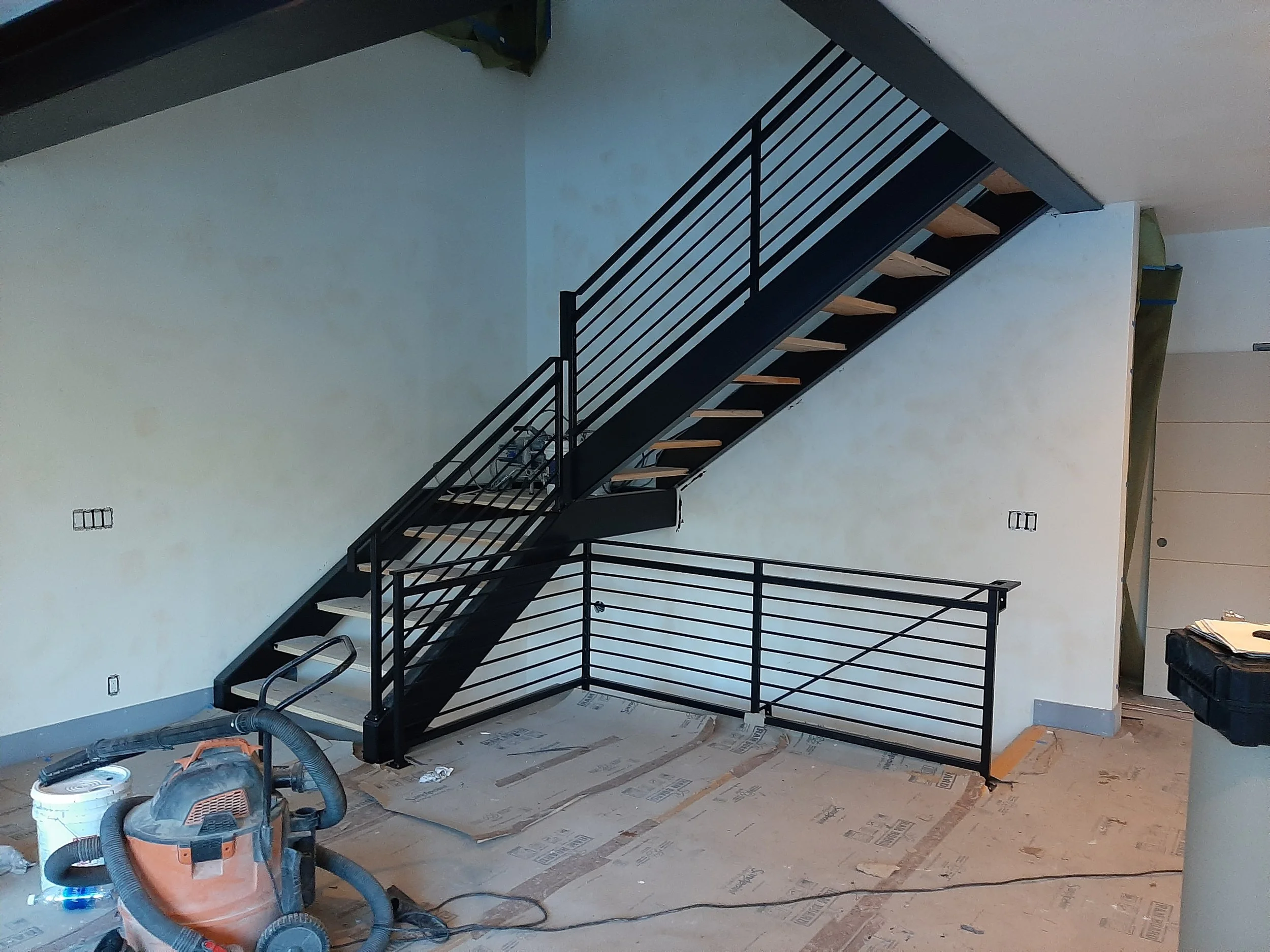 Interior view of a modern staircase with black metal railing and wooden steps, under construction in a room with unfinished floor and walls.