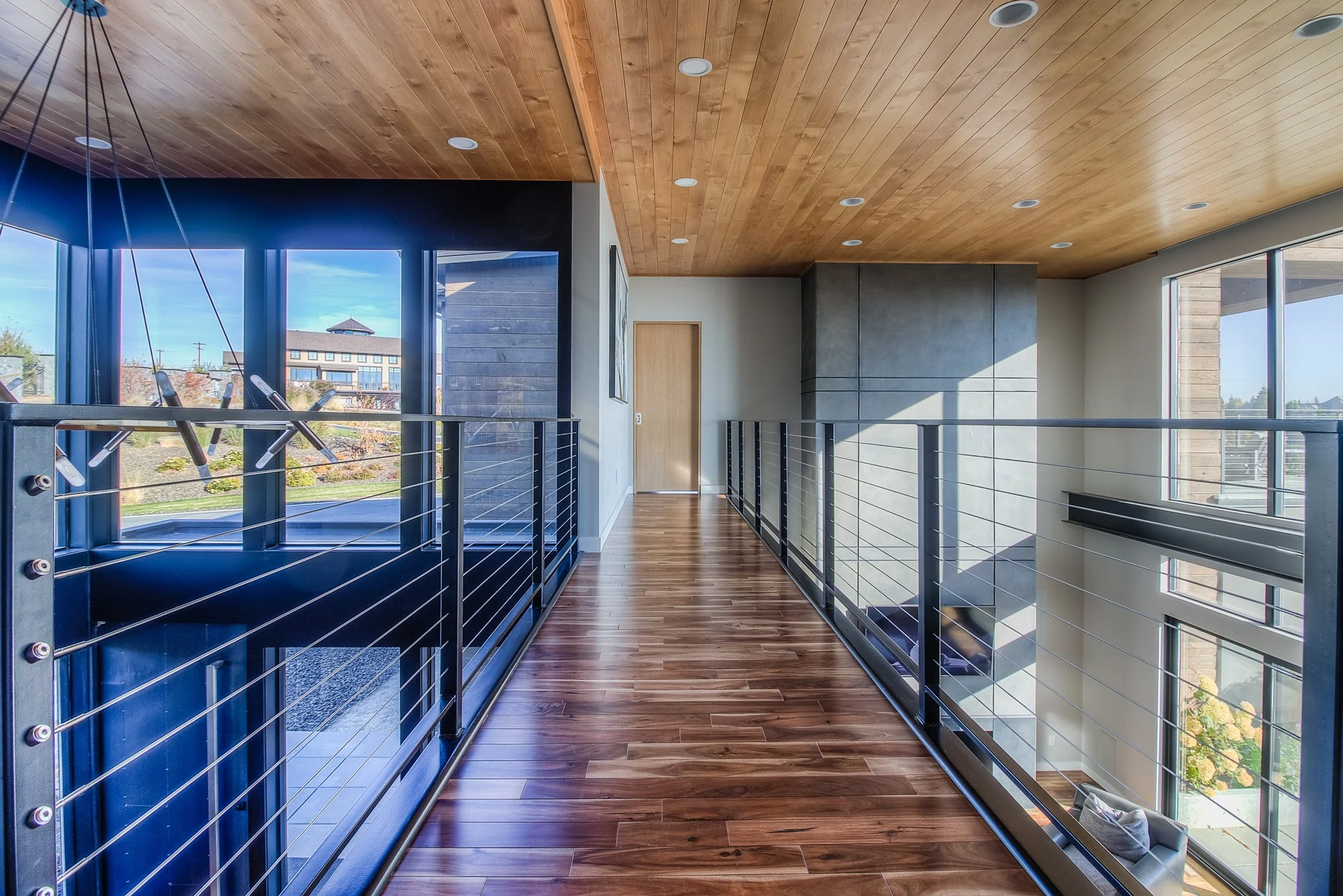 Modern interior hallway with wooden ceiling and floors, metal railing, large windows, and view of neighboring buildings and garden.
