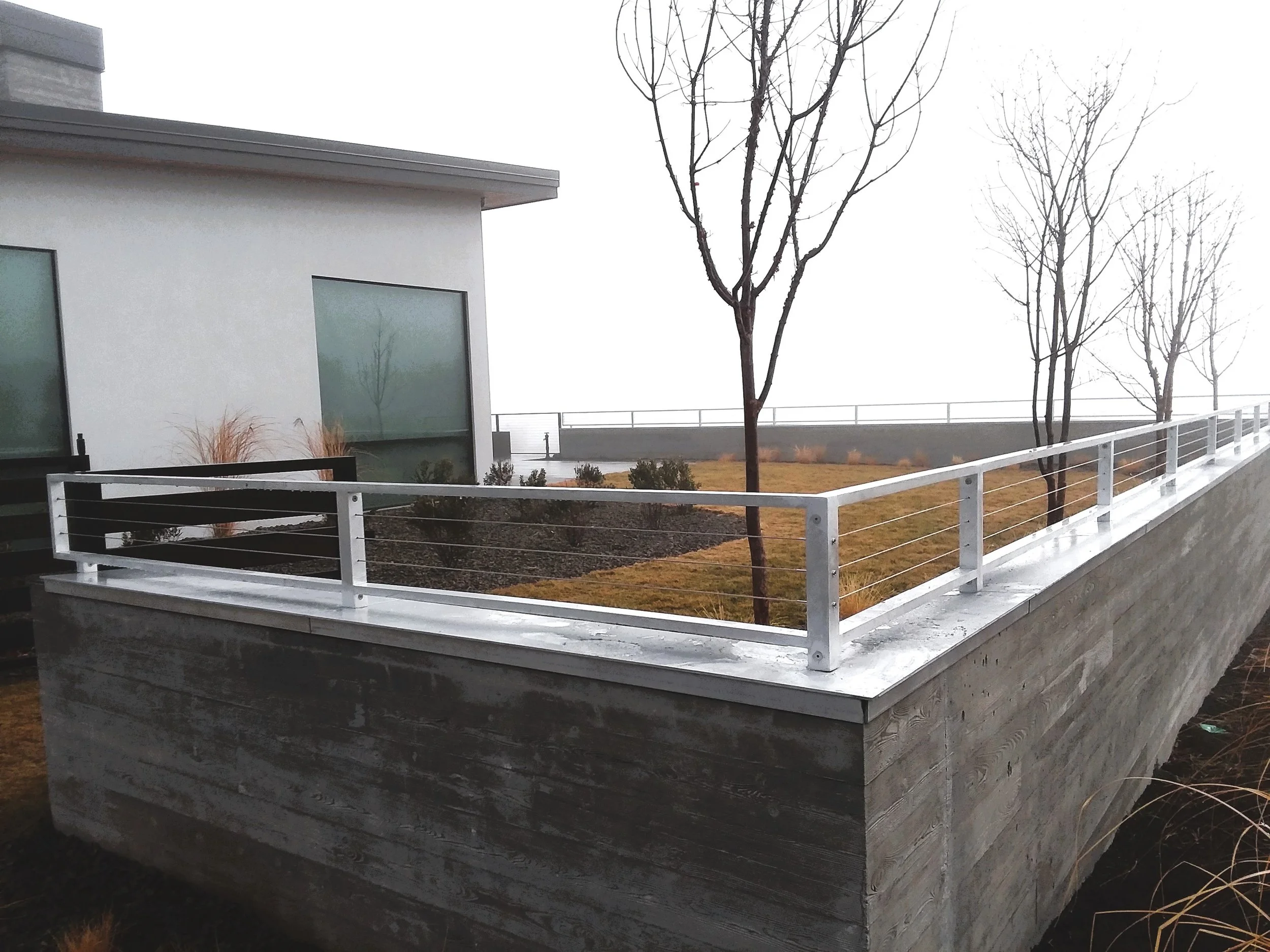 A modern house with large frosted windows, a terraced garden with leafless trees, and a white metal railing on a concrete ledge, in foggy weather.