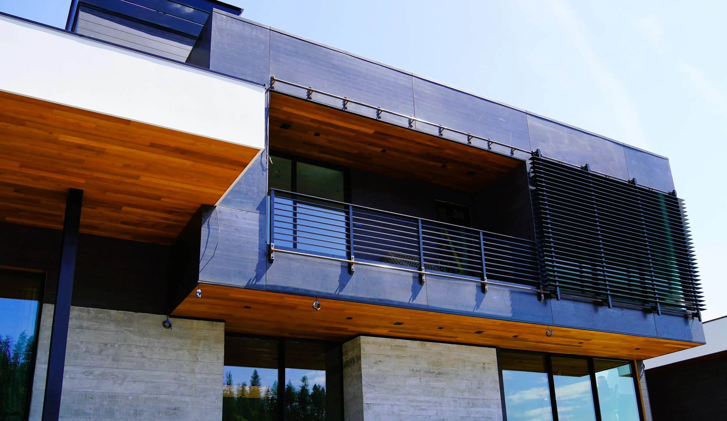 Modern multi-story building with large windows, a balcony with black railing, wooden accents, black louvered panels, and a concrete facade.