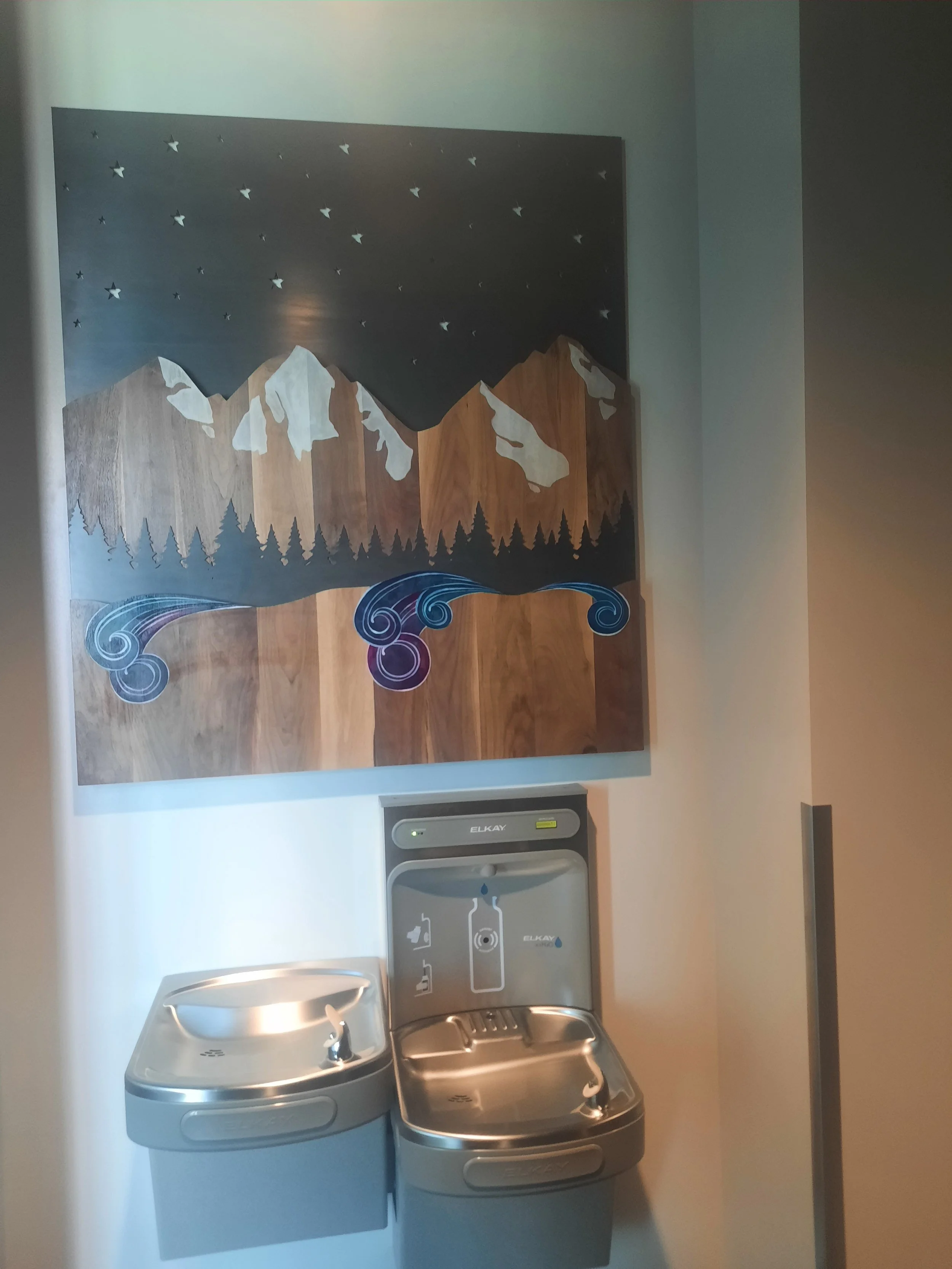 A wooden mountain landscape art piece with stars and swirling water designs hangs above a drinking fountain in a public space.