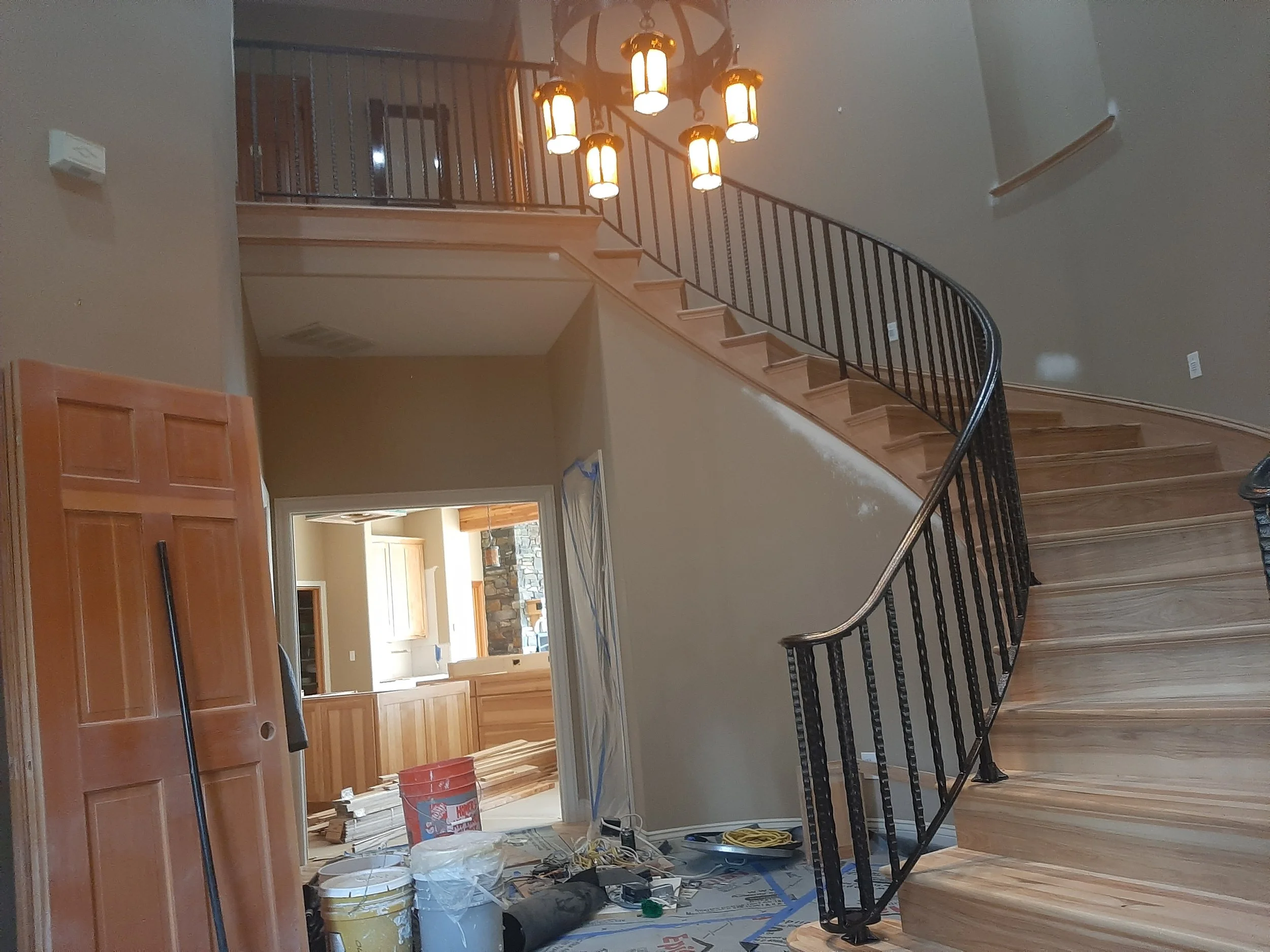 Interior of a house under construction, showing a spiral staircase made of wood and metal handrails, with construction tools and supplies on the floor near a wooden door and open room in the background.
