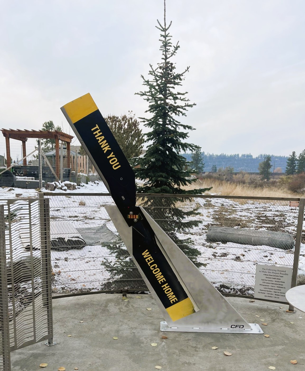 A large outdoor sign resembling a baseball bat with the words 'Thank You' and 'Welcome Home' written on it, mounted on a metal base near a fence and snow-covered ground, with a tree and open field in the background.