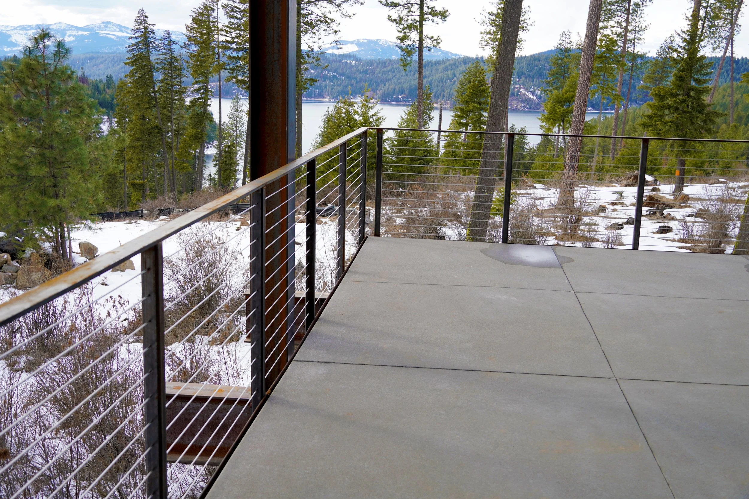A concrete balcony with a metal railing overlooking a snowy landscape with trees, a lake, and mountains in the background.