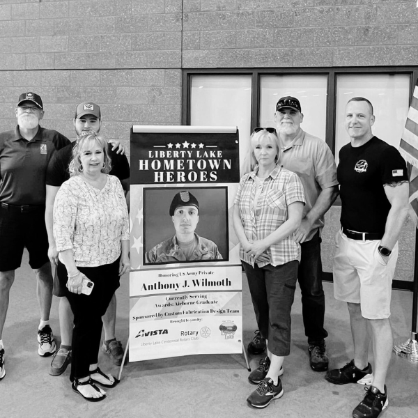 Group of six people standing next to a display honoring Army Private Anthony J. Wilmoth, with a large photograph of him in uniform, inside a building.