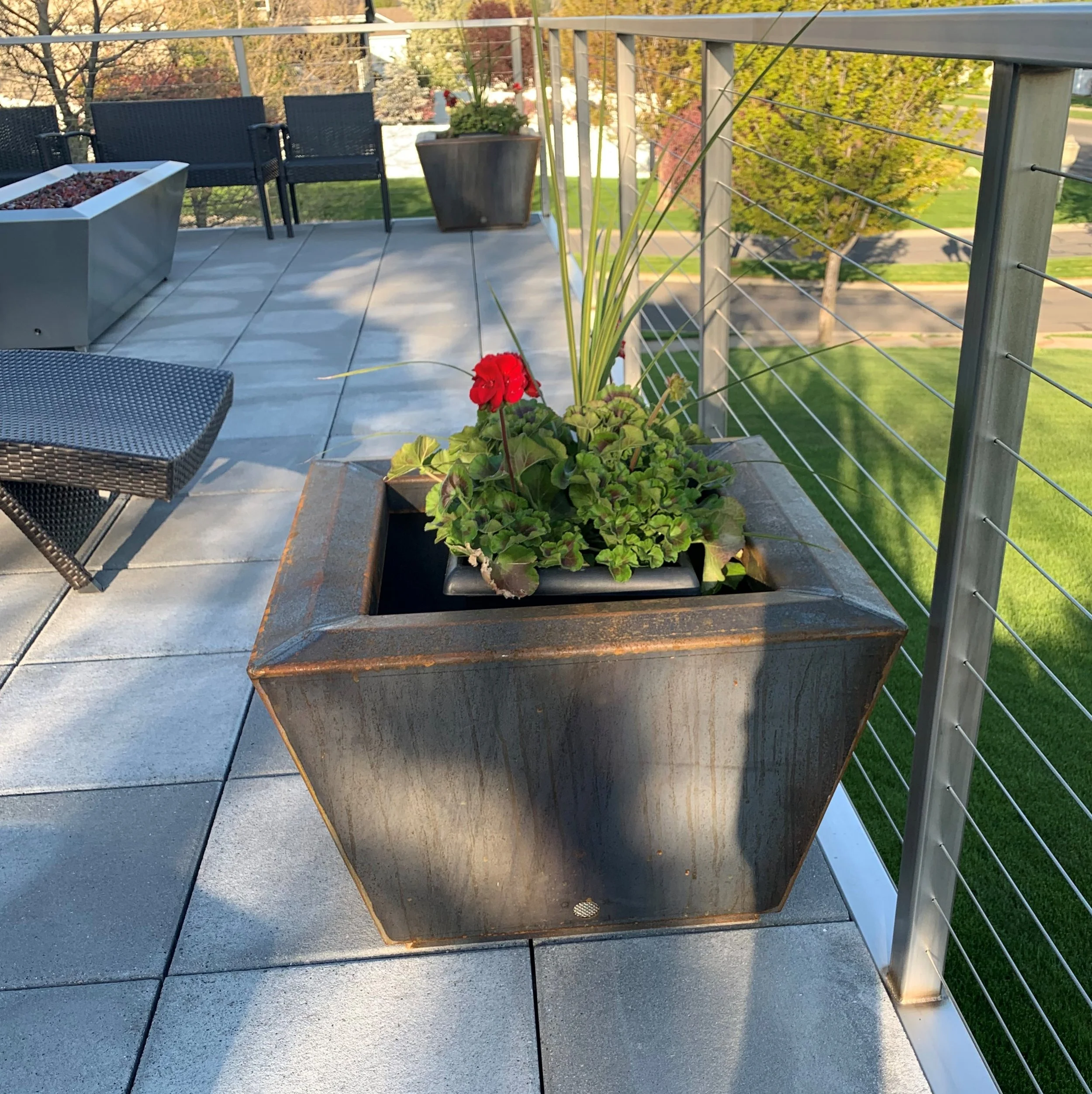 A large, rustic, square metal planter box with a rusted finish, containing green leafy plants and a single red flower, on a patio with gray tiles and a metal railing overlooking a grassy area with trees.