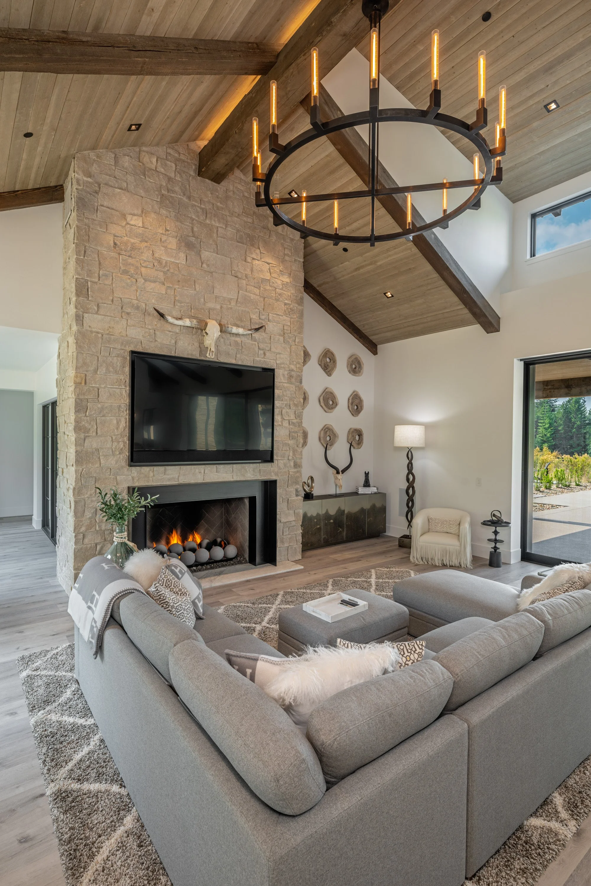 Living room with a stone fireplace, mounted television, modern decor, and a large sectional sofa arranged around a coffee table. The ceiling has wooden beams and a modern chandelier. There's a floor lamp, a small white chair, and sliding glass doors 