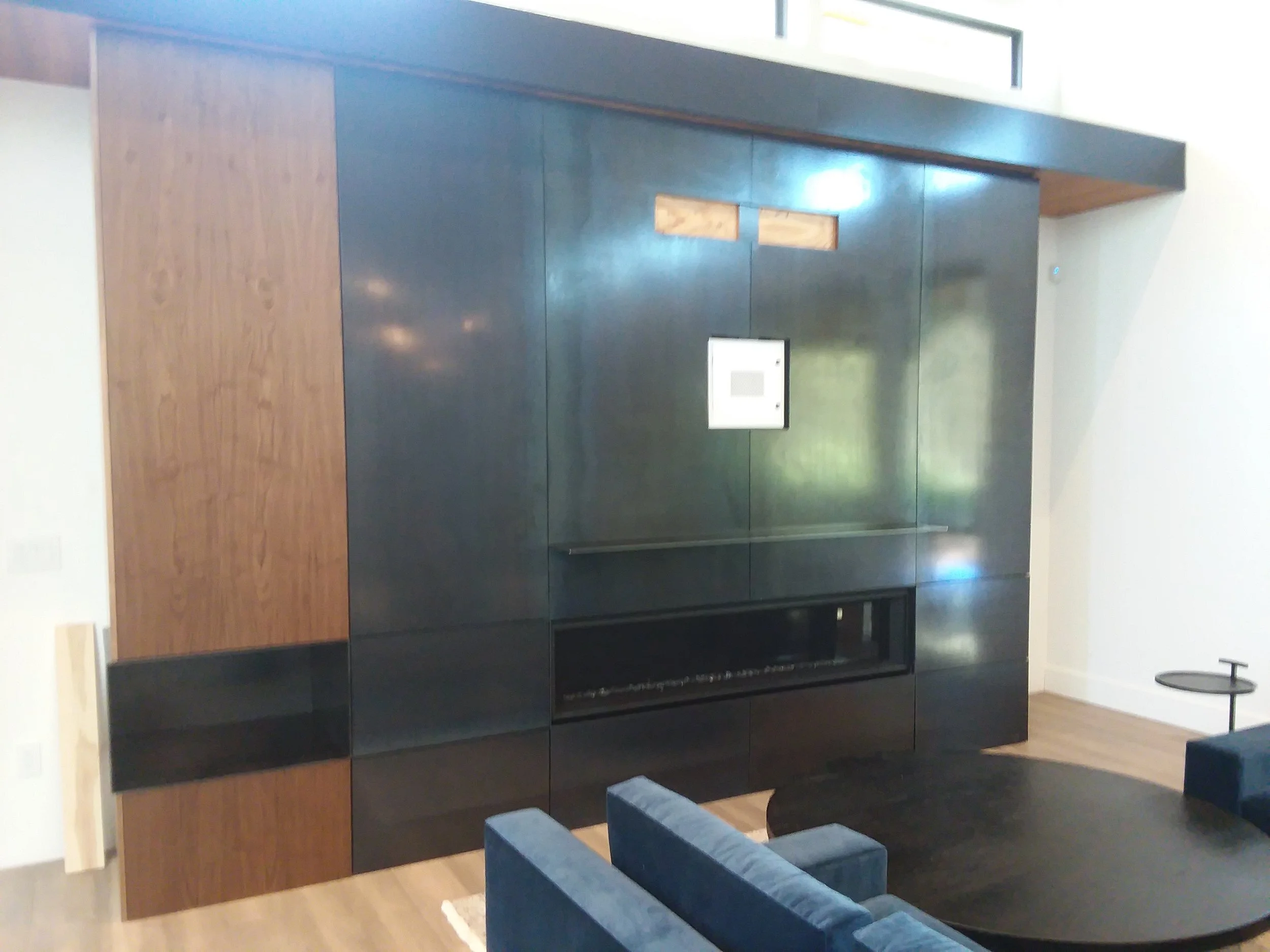 Modern living room with dark wood and black built-in wall unit, a small digital device mounted on the wall, blue sofa, and a black round coffee table, with hardwood floors and a white wall.