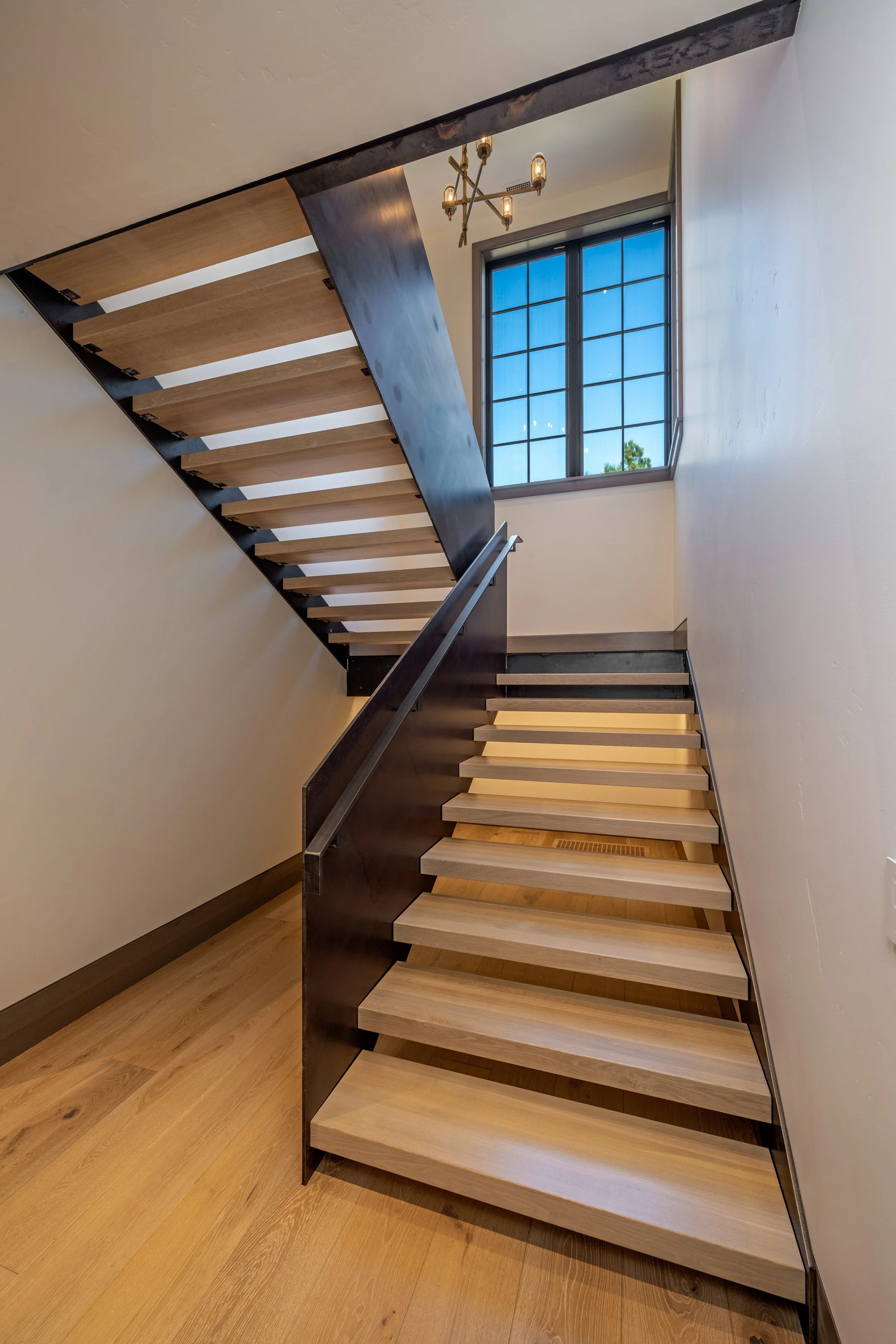 Interior view of a modern wooden staircase with black metal supports, leading to an upper floor near a window with a grid pattern, under a modern chandelier, with light-colored walls and wooden flooring.