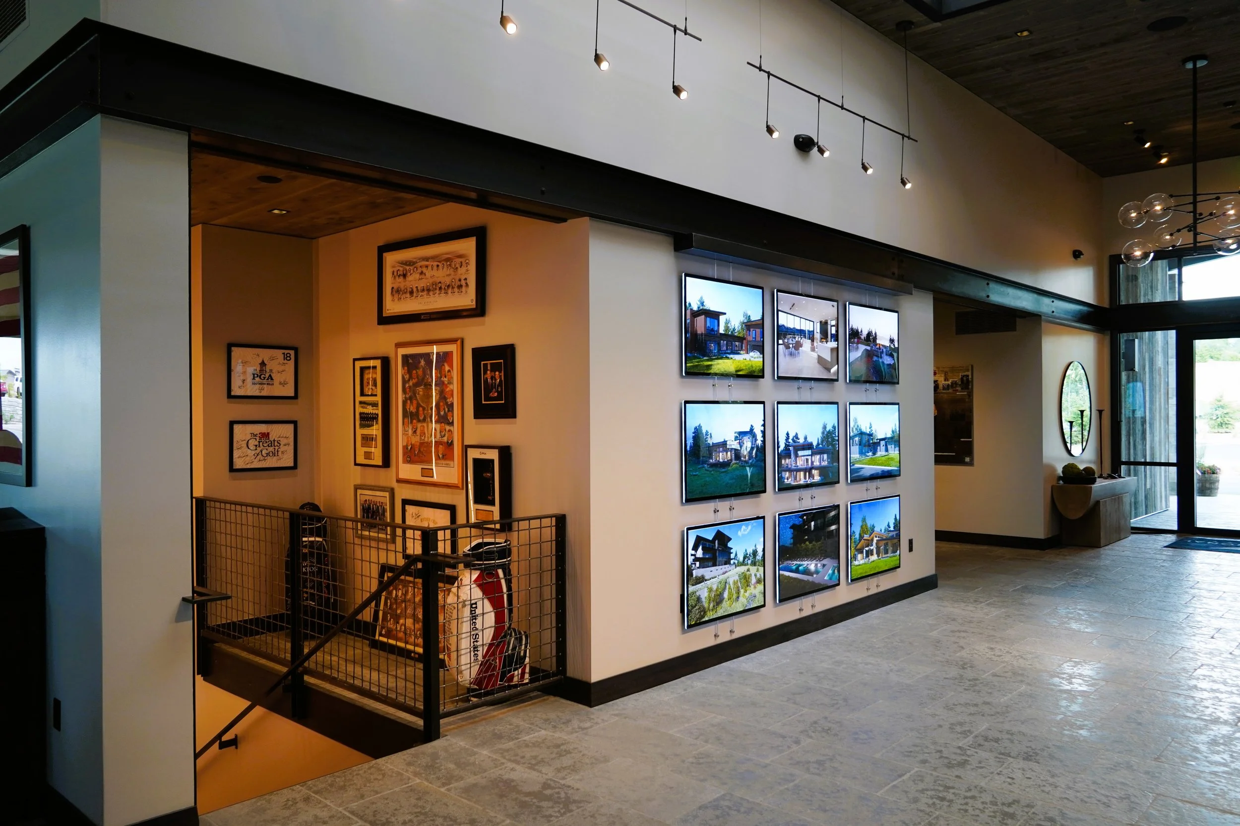 Interior of a modern building with framed photographs and artwork on the walls, a display of mounted photos or videos, a staircase with golf clubs, and an entrance door leading outside.