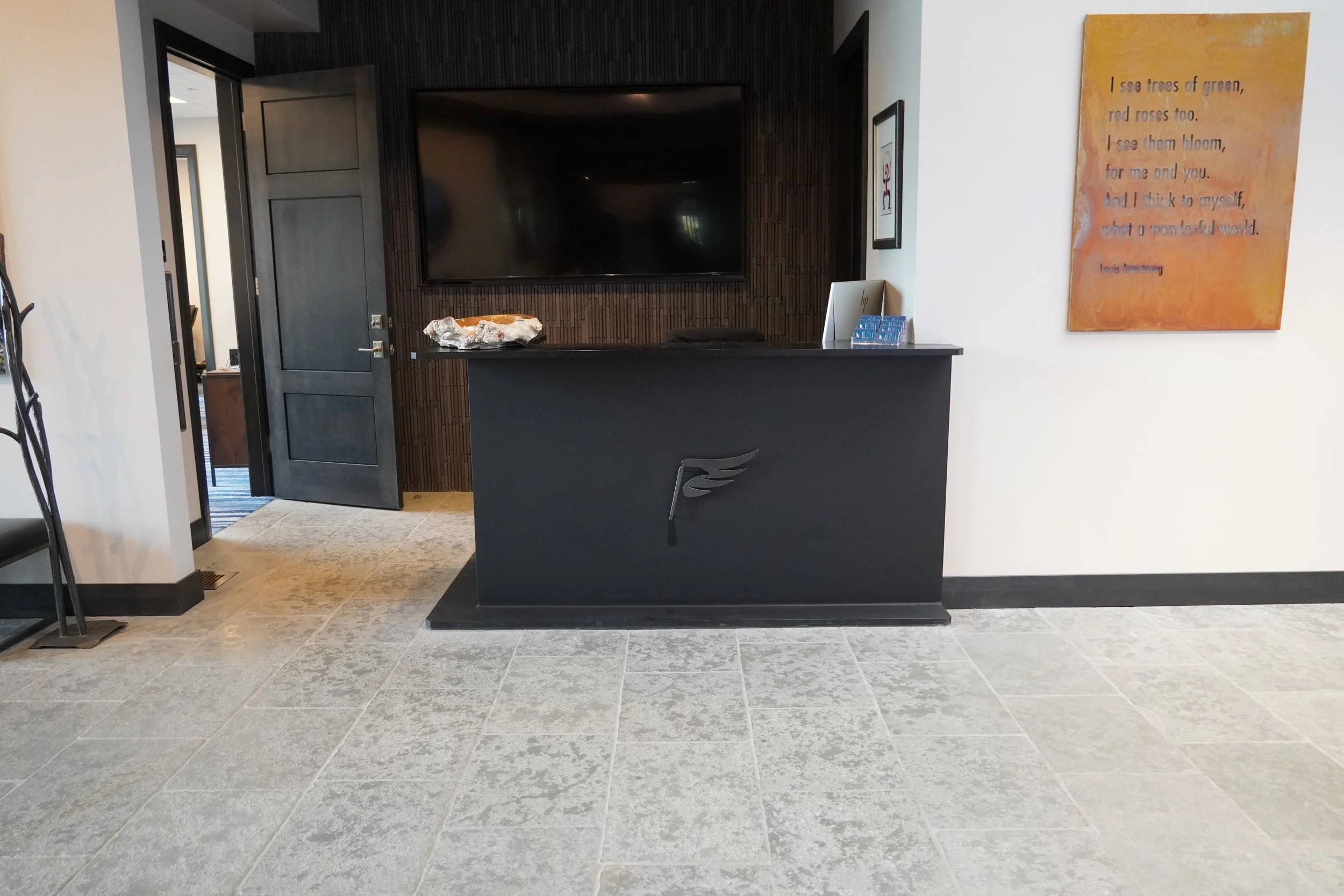 Hotel lobby reception desk with a black front panel displaying a logo, a computer, a brochure holder, and items on top, against a dark wood accent wall with a large flat-screen TV. To the right, a wall features a colorful painting with a quote, and t