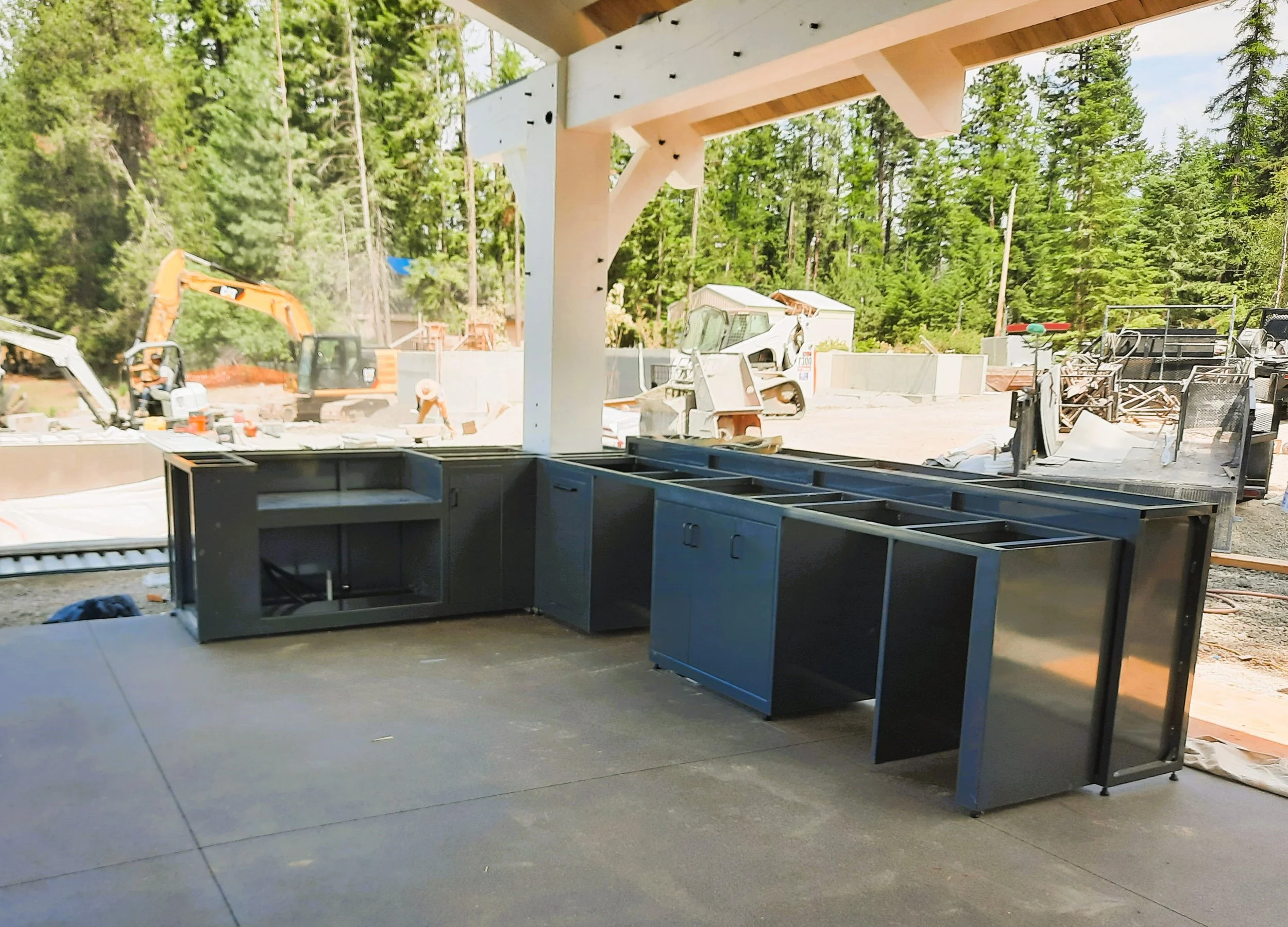 Construction site with black kitchen cabinets installed, construction machinery, and workers in the background, surrounded by trees.