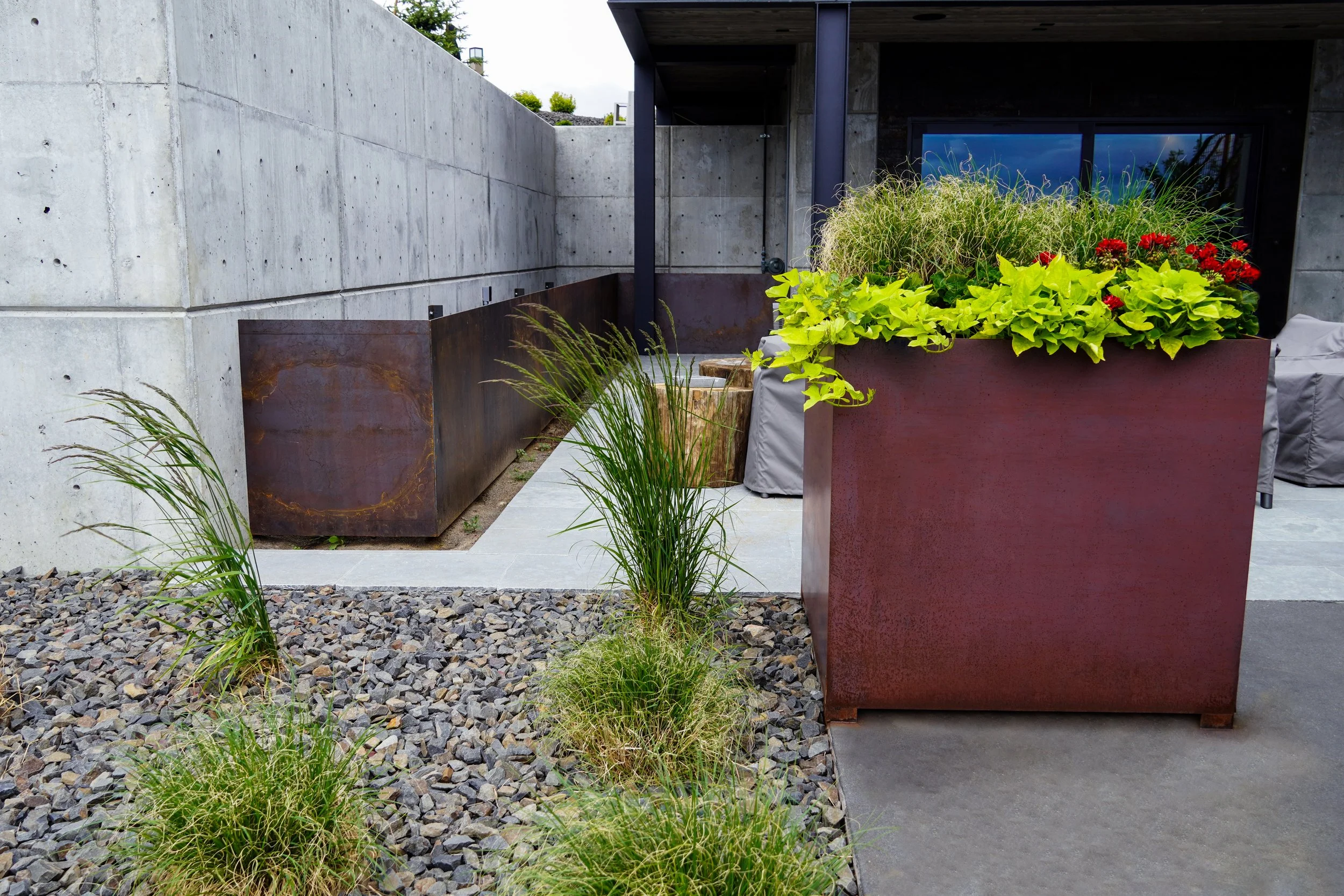 Modern outdoor patio with concrete walls, large rust-colored metal planters, green plants, and a gray patio cover.