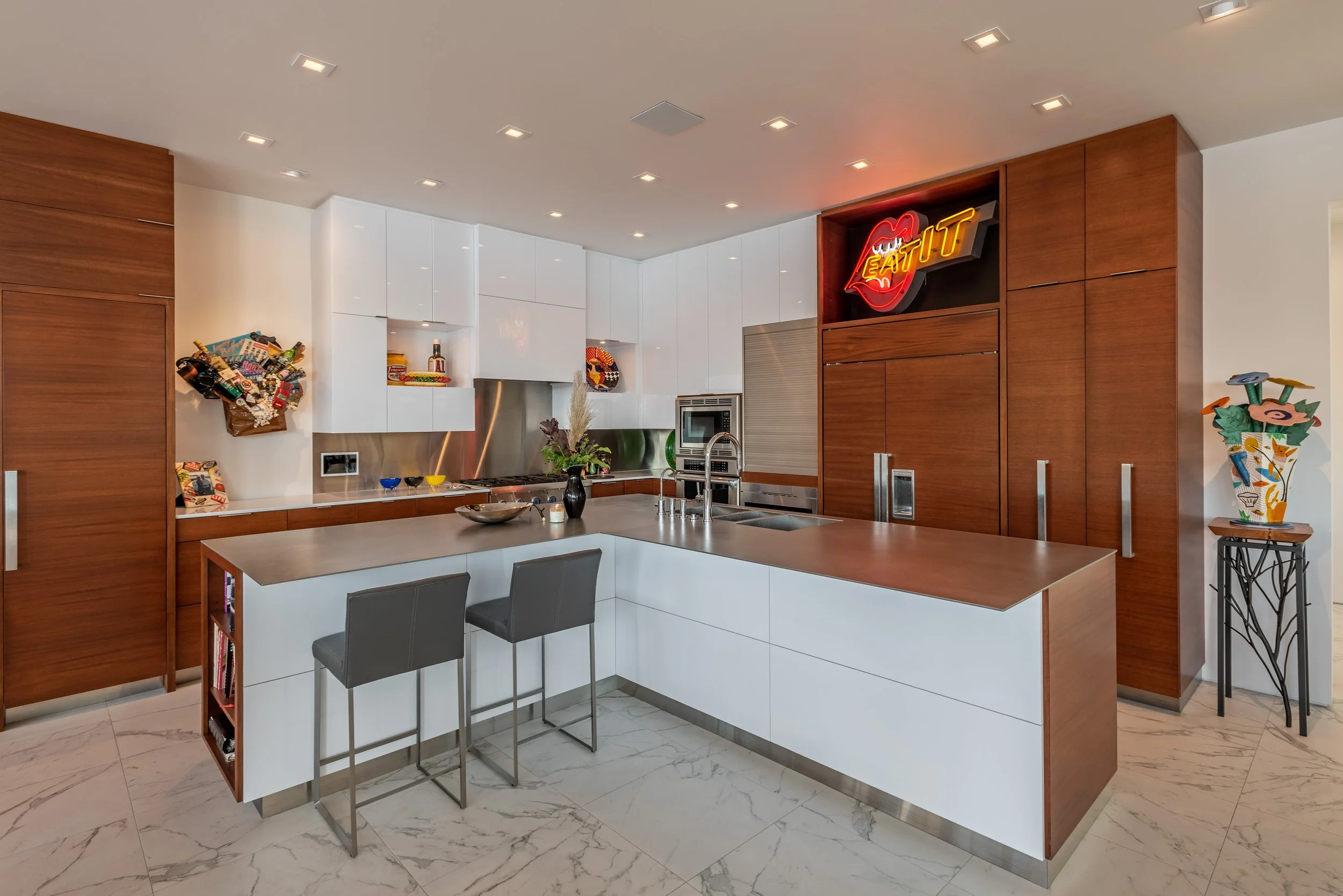Modern kitchen with white and wood cabinetry, marble floor, and a kitchen island with two gray barstools. Decor includes a black vase with flowers, colorful art pieces, and a neon sign on the wall reading 'EAT' in red and yellow.
