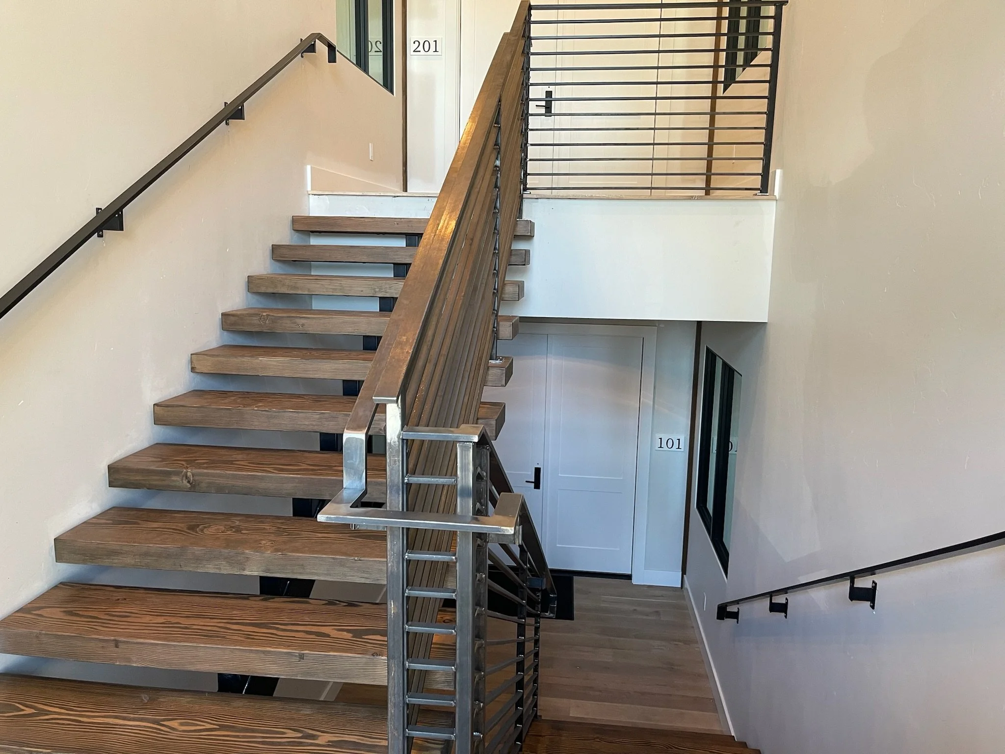 Interior view of a multi-floor stairwell with wooden stairs, metal handrails, and numbered apartment doors visible at the top and bottom of the stairwell.