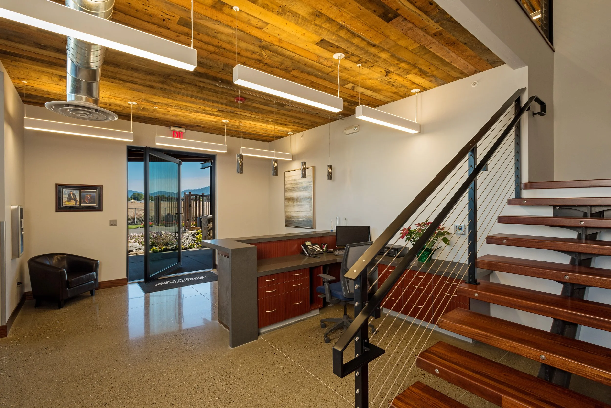 Office reception area with a wooden ceiling, a glass door leading outside, a reception desk with a computer and telephone, a small armchair, and a staircase with wooden steps and a metal railing.