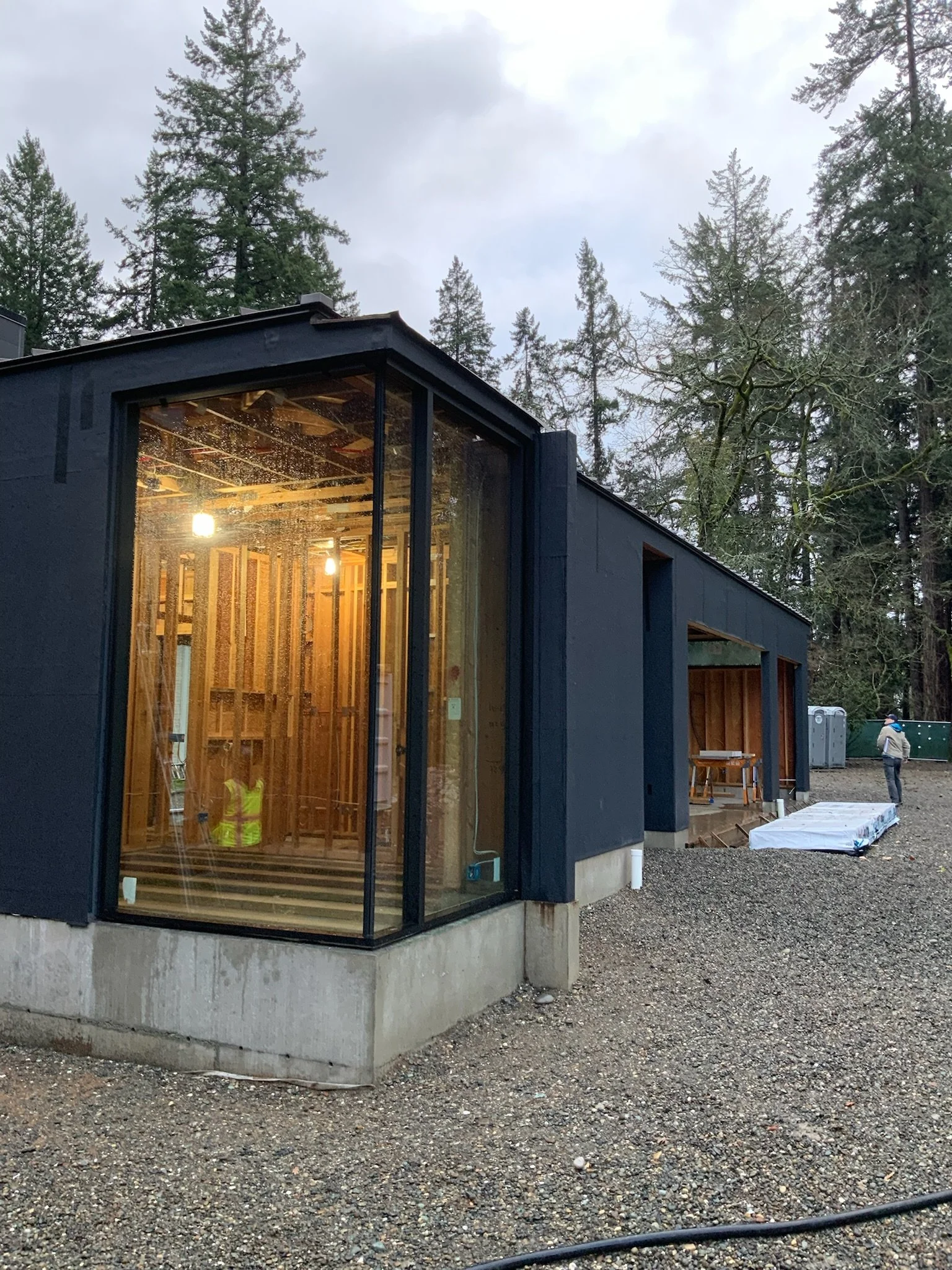 Construction of a modern building with large glass windows in a wooded area, showing the interior framework and workers on site.