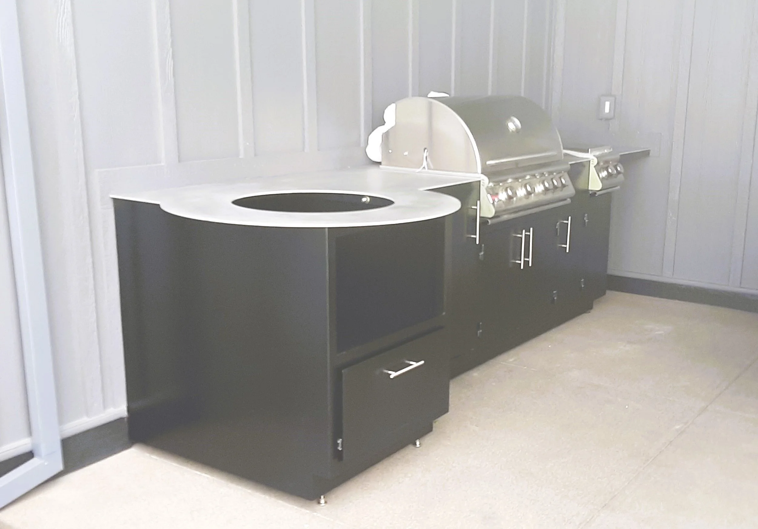An outdoor kitchen setup with a round black countertop grill and a stainless steel barbecue grill on a black cabinet against a gray paneled wall.