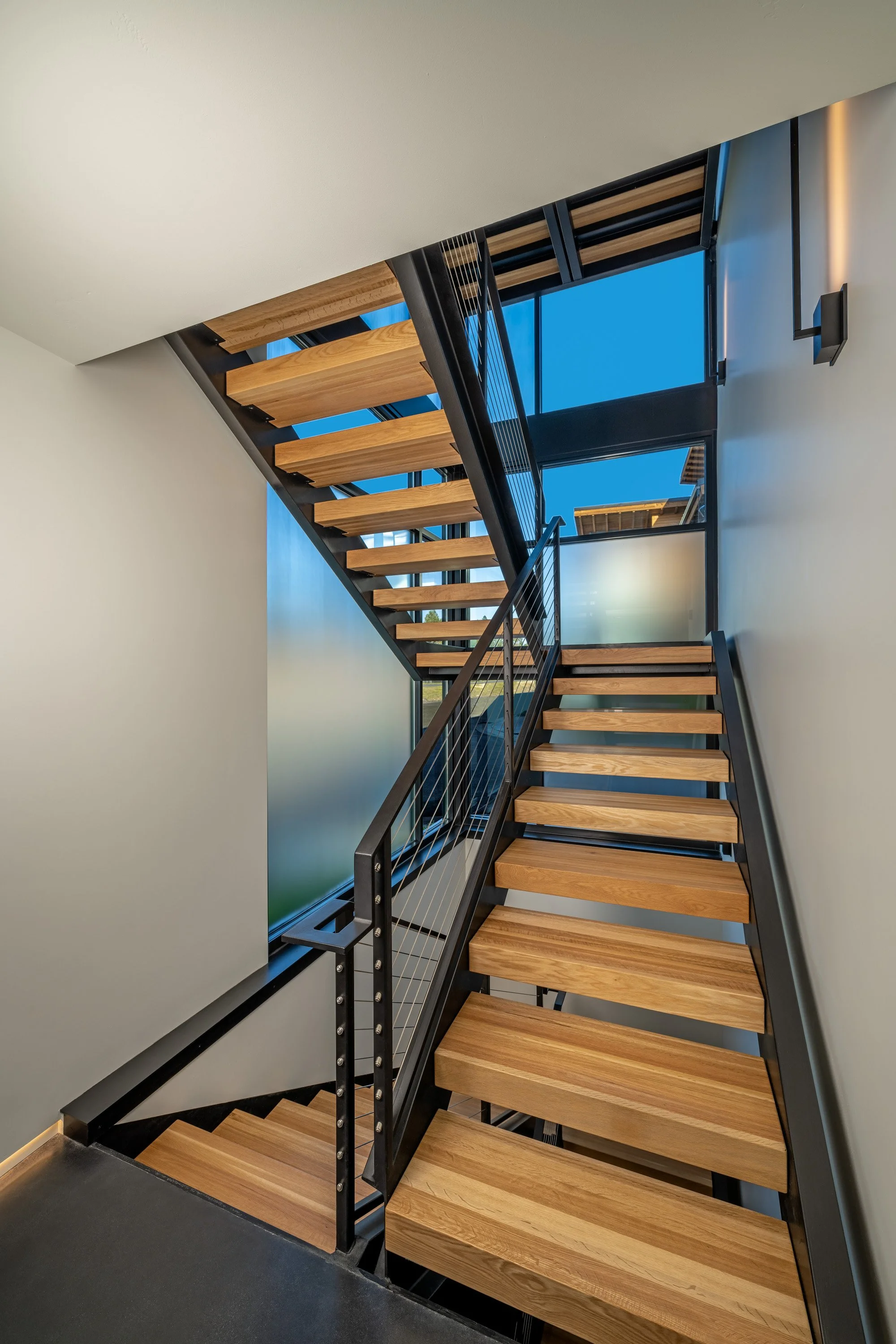 Interior view of a modern staircase with wooden steps and black metal railings, next to large windows showing a blue sky.