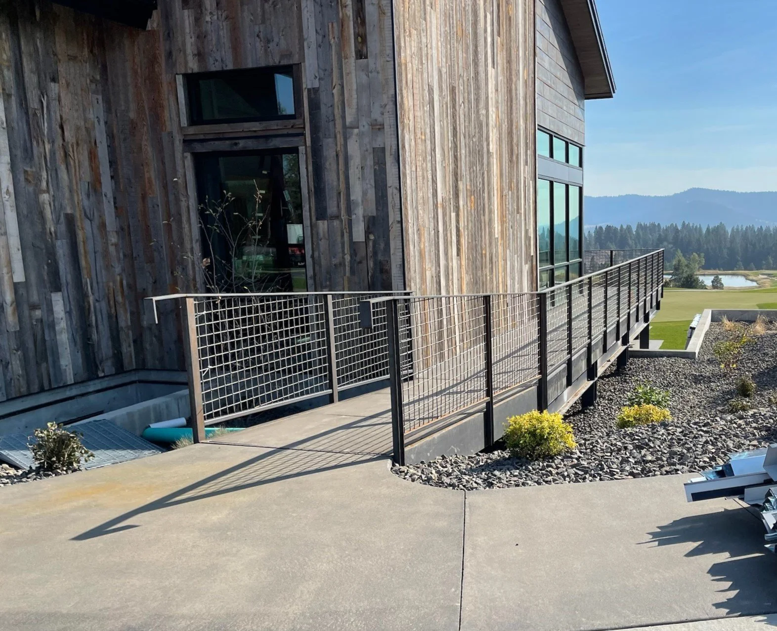 Modern building with wooden exterior, large windows, and a metal railing pathway, overlooking a scenic landscape with mountains, fields, and water.