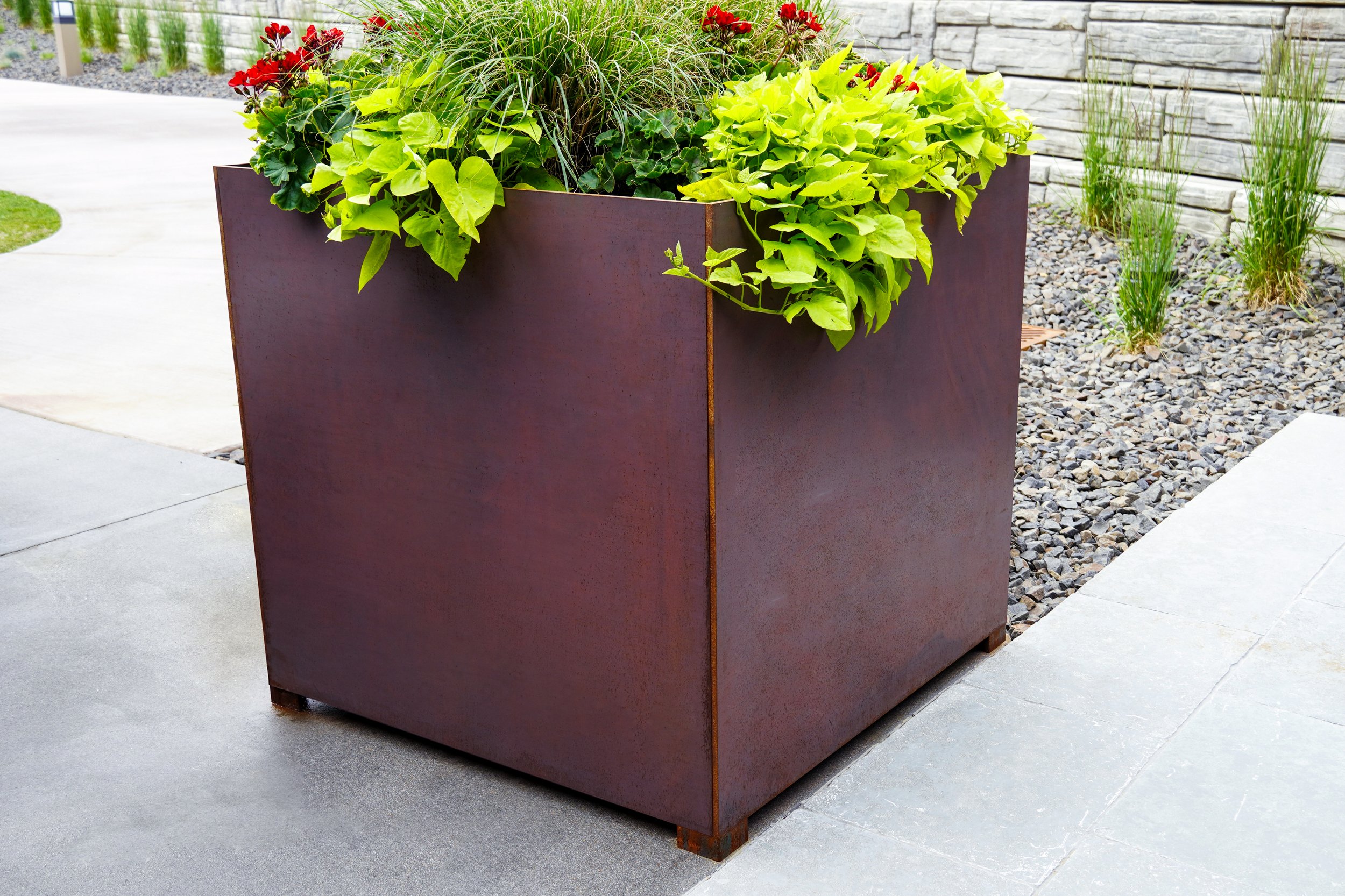 A large, dark rust-colored metal planter box filled with green leafy plants and red flowers, situated on a concrete patio near a stone wall and landscaped area.