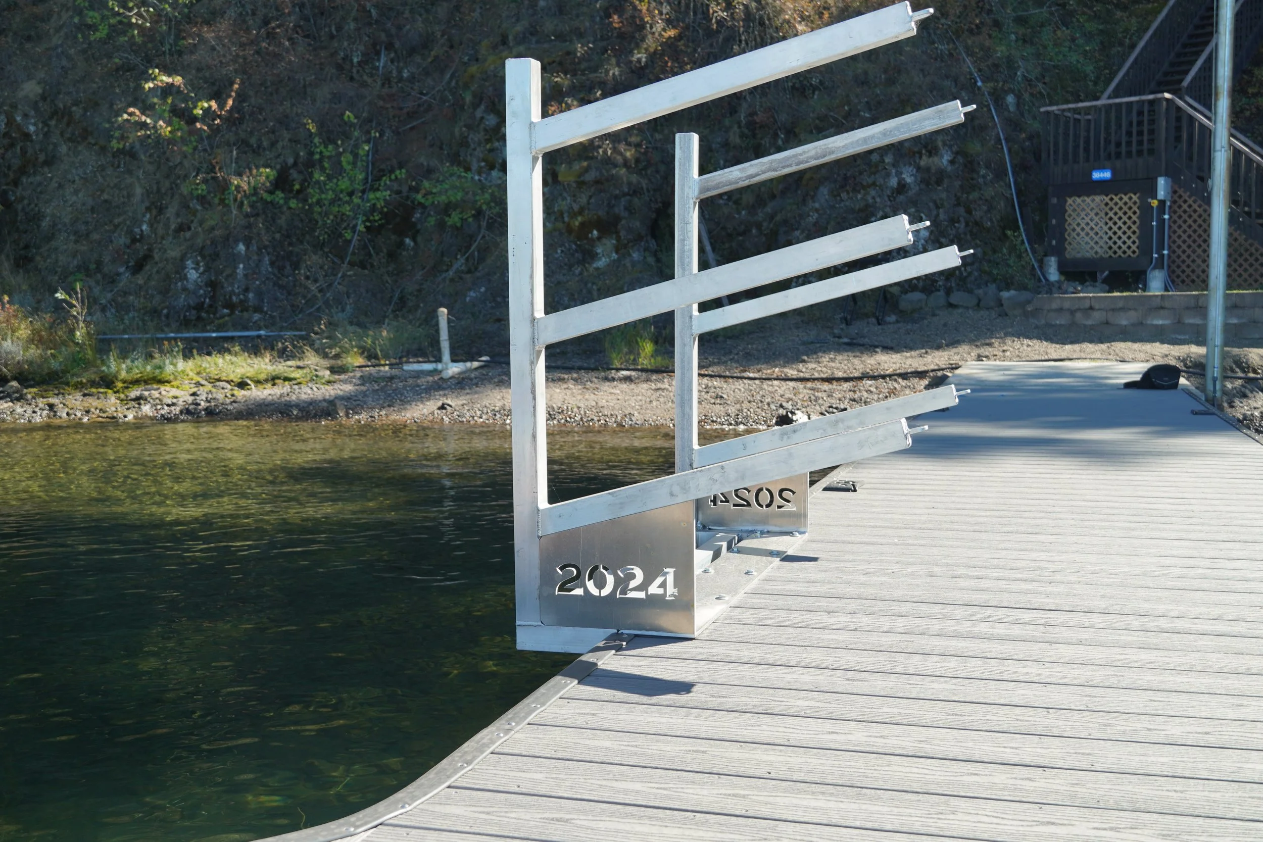 Empty dock with a metal railing and the year 2024 inscribed on it, extending over calm water with a wooded hillside in the background.