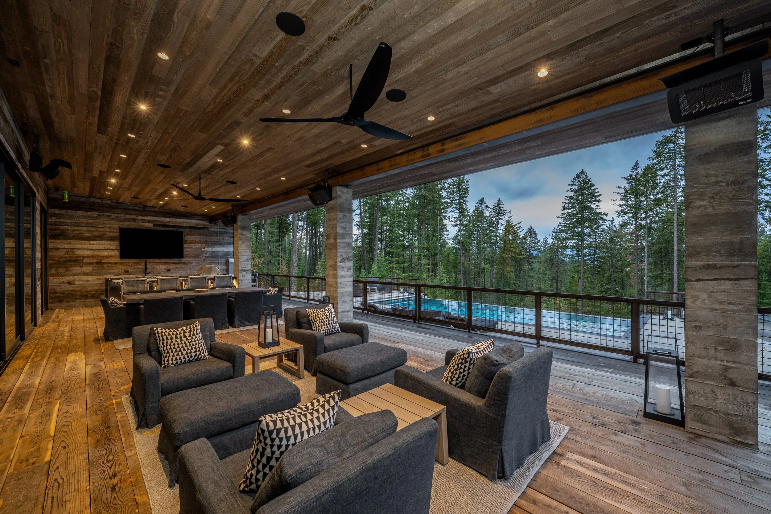 Indoor lounge area with wooden walls and ceiling, modern black ceiling fans, leather armchairs with patterned pillows, wooden tables, and a large open space overlooking a forest with a pool.