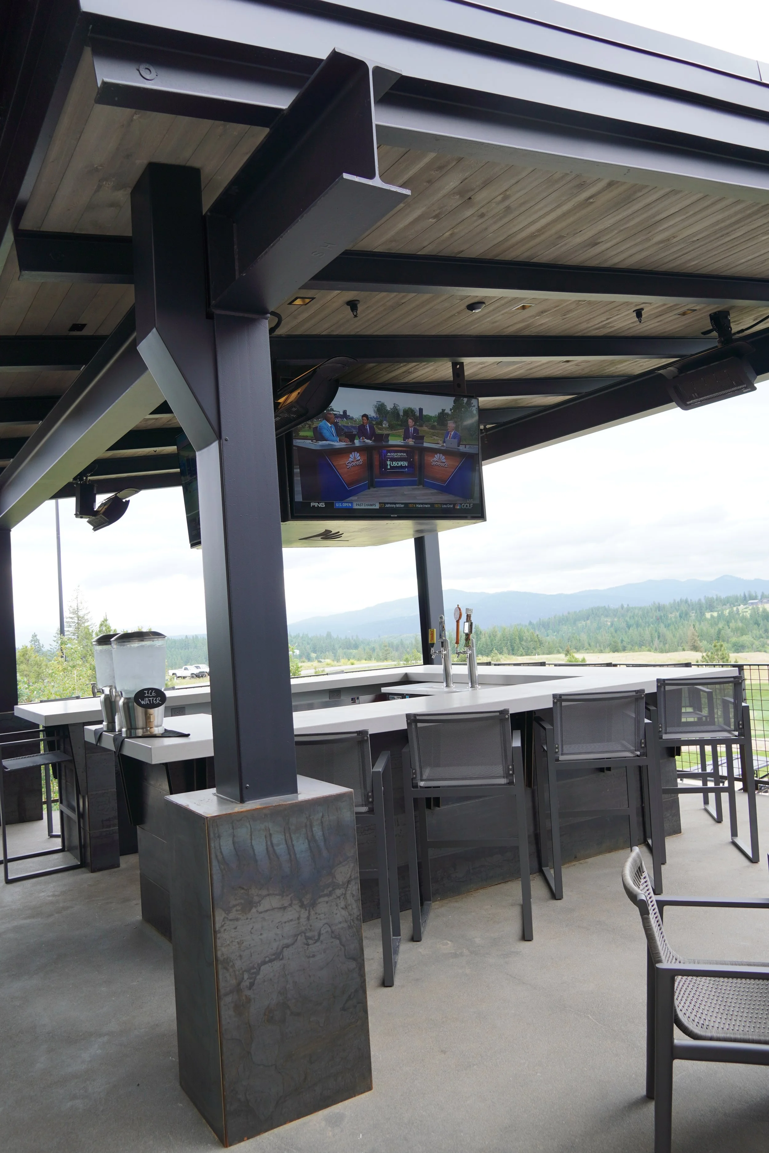 An outdoor bar area with a white countertop, bar stools, a TV screen showing a sports broadcast, and a scenic mountainous landscape in the background.