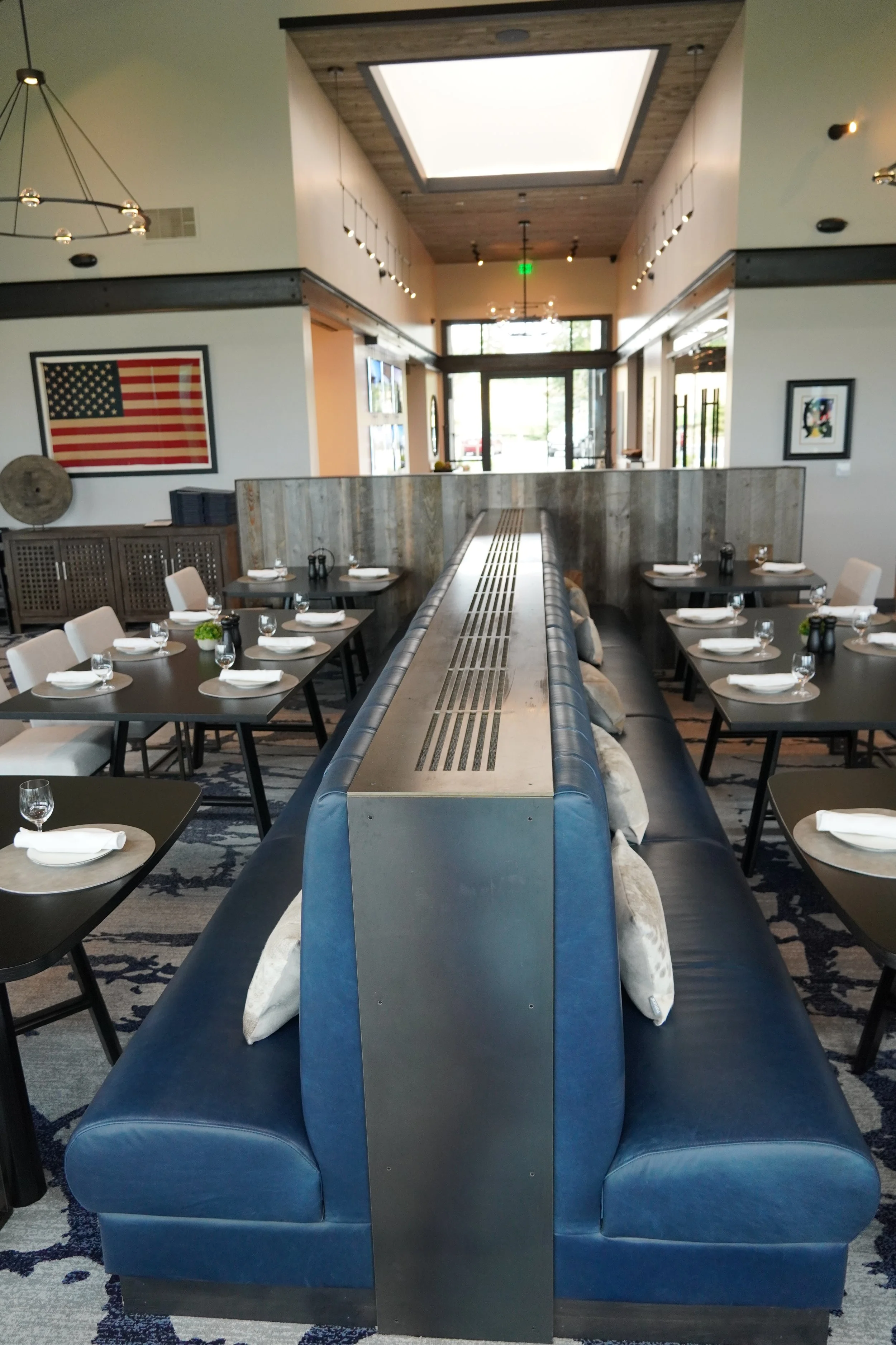 Inside a restaurant, view of a blue booth with pillows, surrounded by tables set with plates, glasses, and napkins, with an American flag on the wall and modern decor.