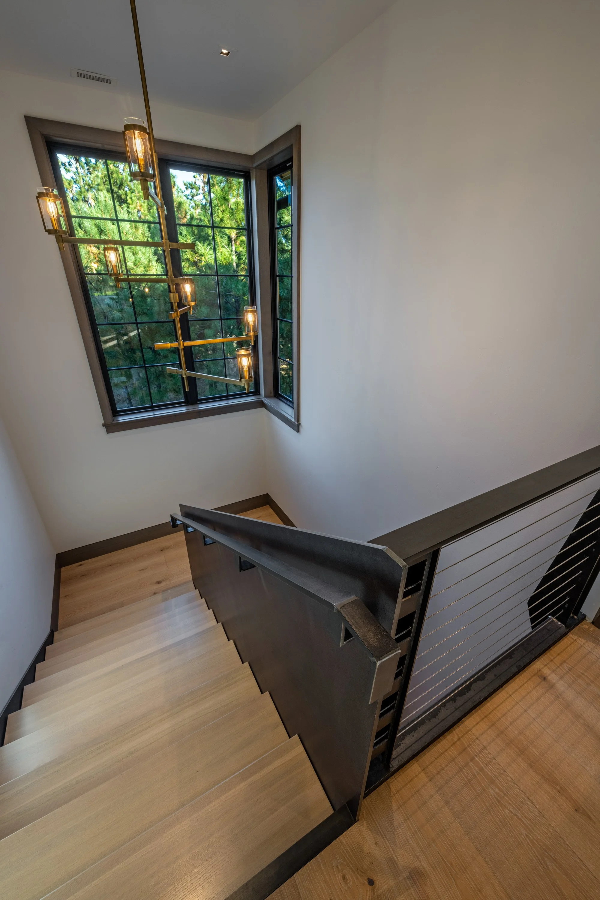 A staircase with wooden steps and metal railing, viewed from above, near a large window with a view of green trees, and a modern hanging chandelier with multiple lights.