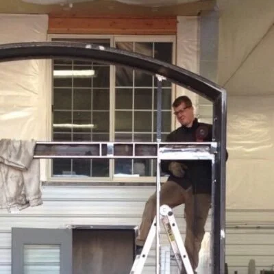 A man working on a large window frame inside a garage, standing on a ladder.