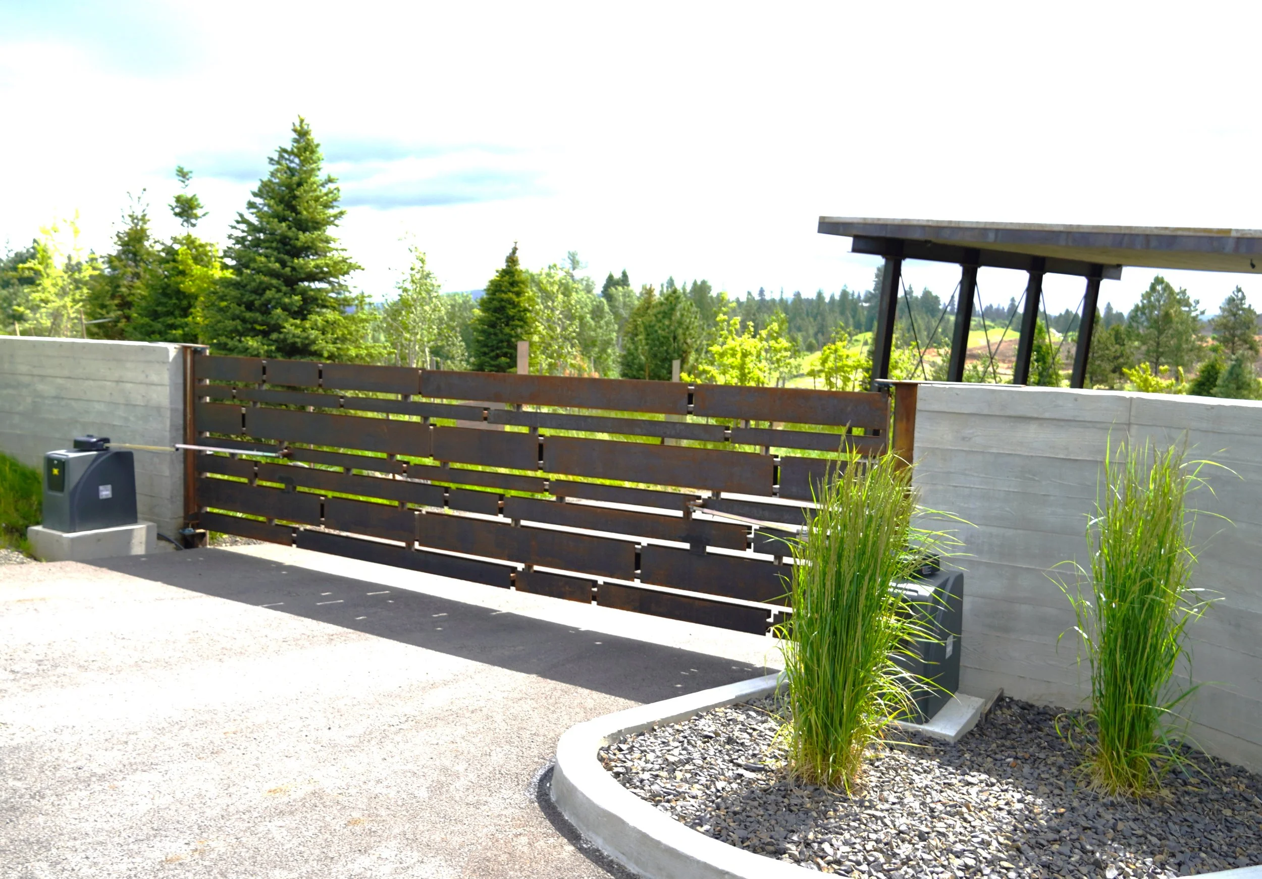 A modern residential driveway with a wooden gate, landscaping with tall grass, and a concrete wall against a backdrop of trees and a partly cloudy sky.