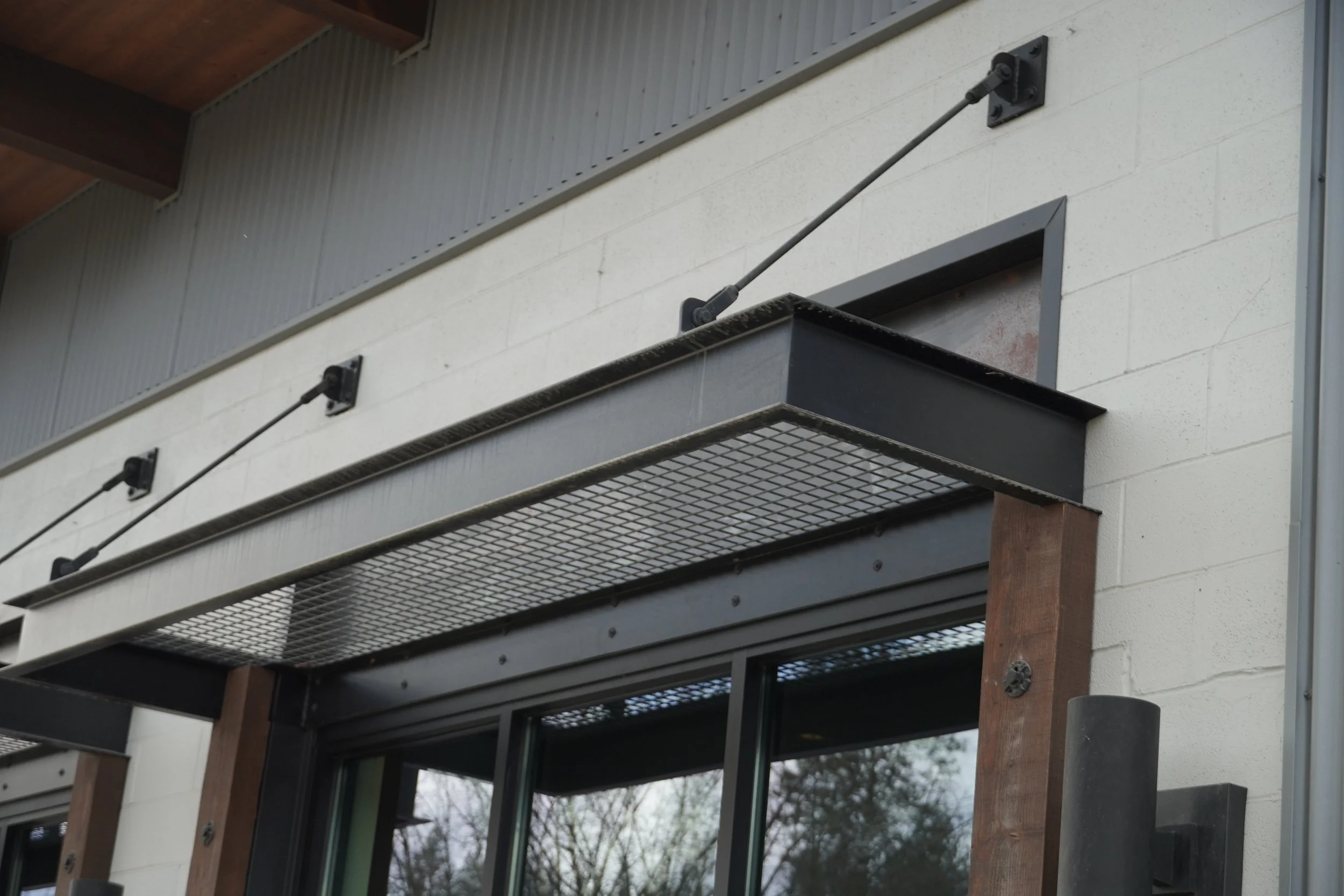 A close-up of a balcony railing with metal mesh and support brackets attached to a light-colored exterior wall of a building, with trees and cloudy sky visible in the background.