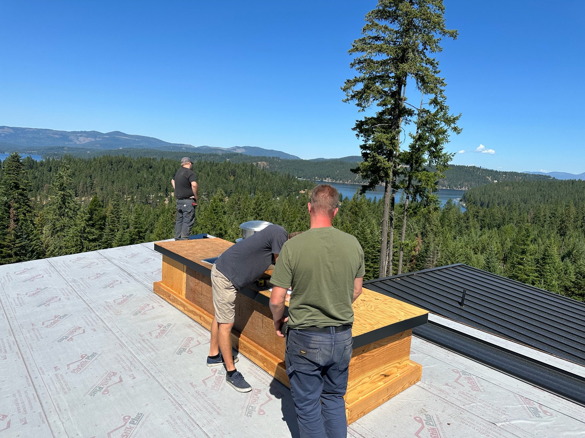 Three men working on a rooftop with a scenic view of a forest, lake, and mountains in the background. One man is standing near the edge, while the other two are near a wooden structure.