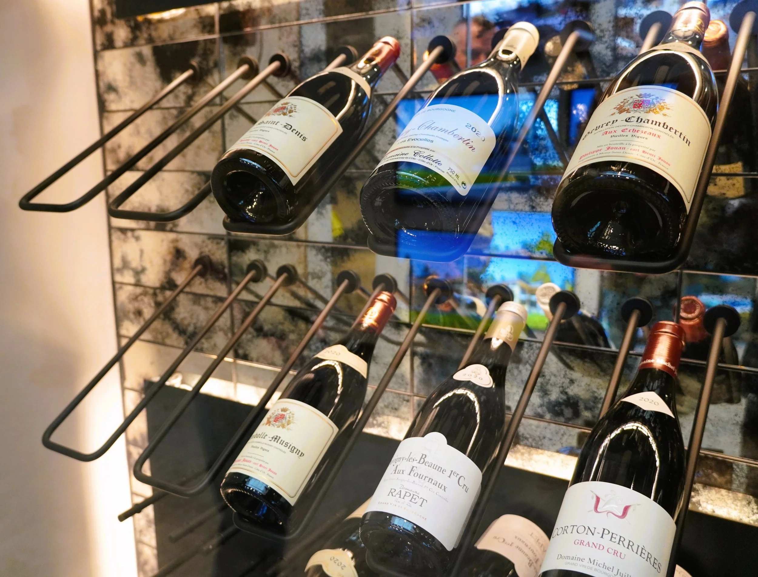 Wine bottles on a metal rack in front of a decorative background that resembles a brick wall. The bottles are of various types and labels.