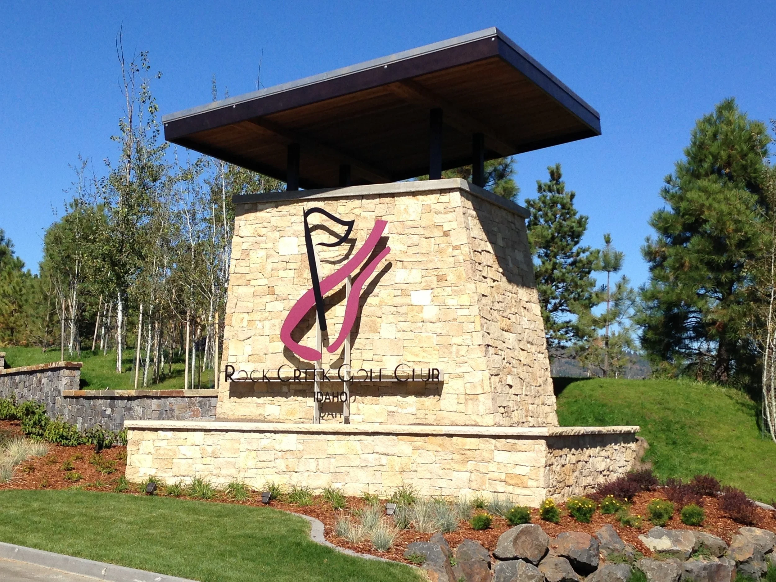 Entrance sign for Rock Creek Golf Club in Idaho, featuring a stylized golf club and ball icon, on a stone structure with landscaping and trees in the background.