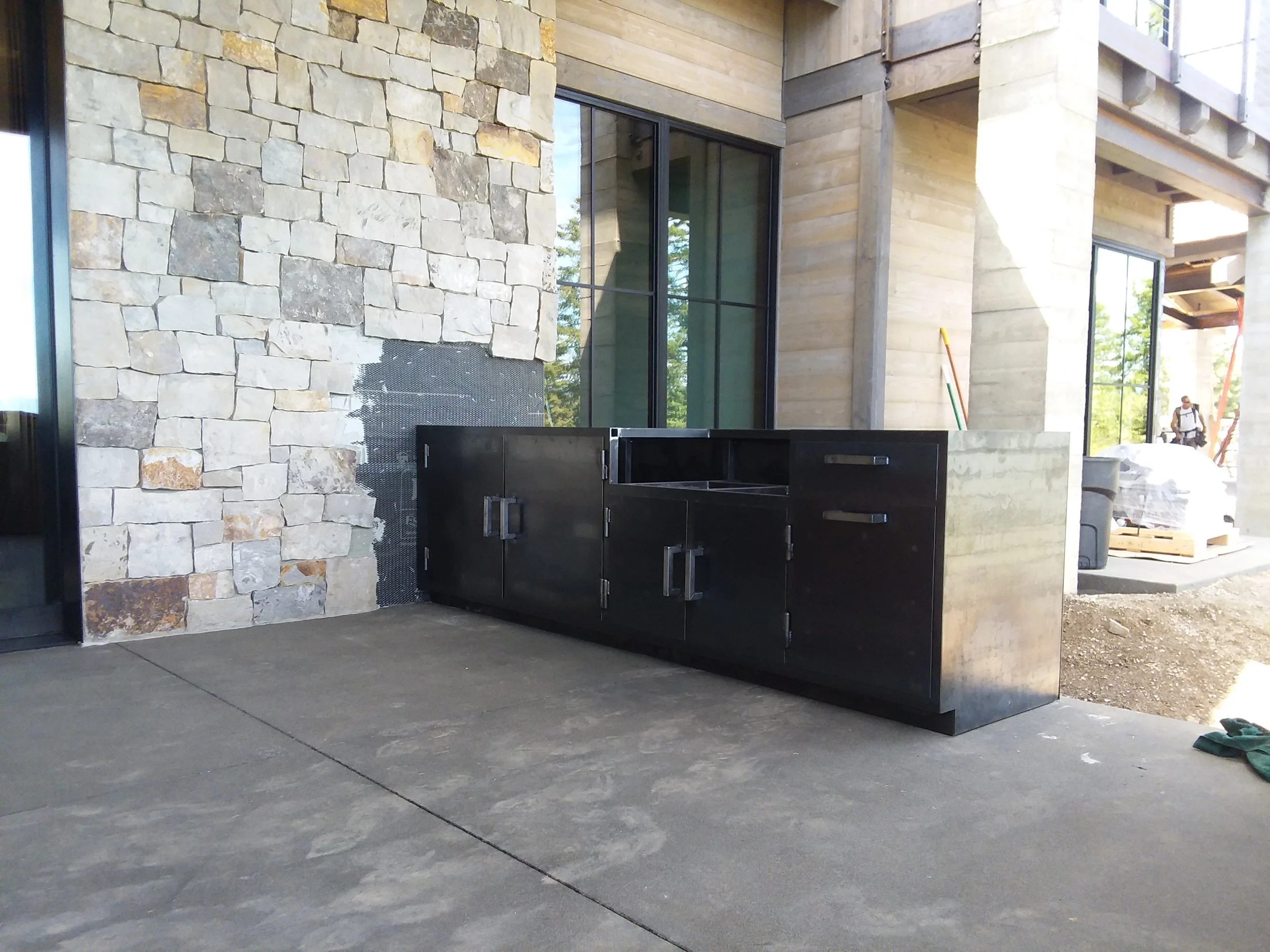 Outdoor kitchen with a black metal cabinet against a beige stone wall, on a concrete patio of a house under construction with large glass windows and wooden beams.